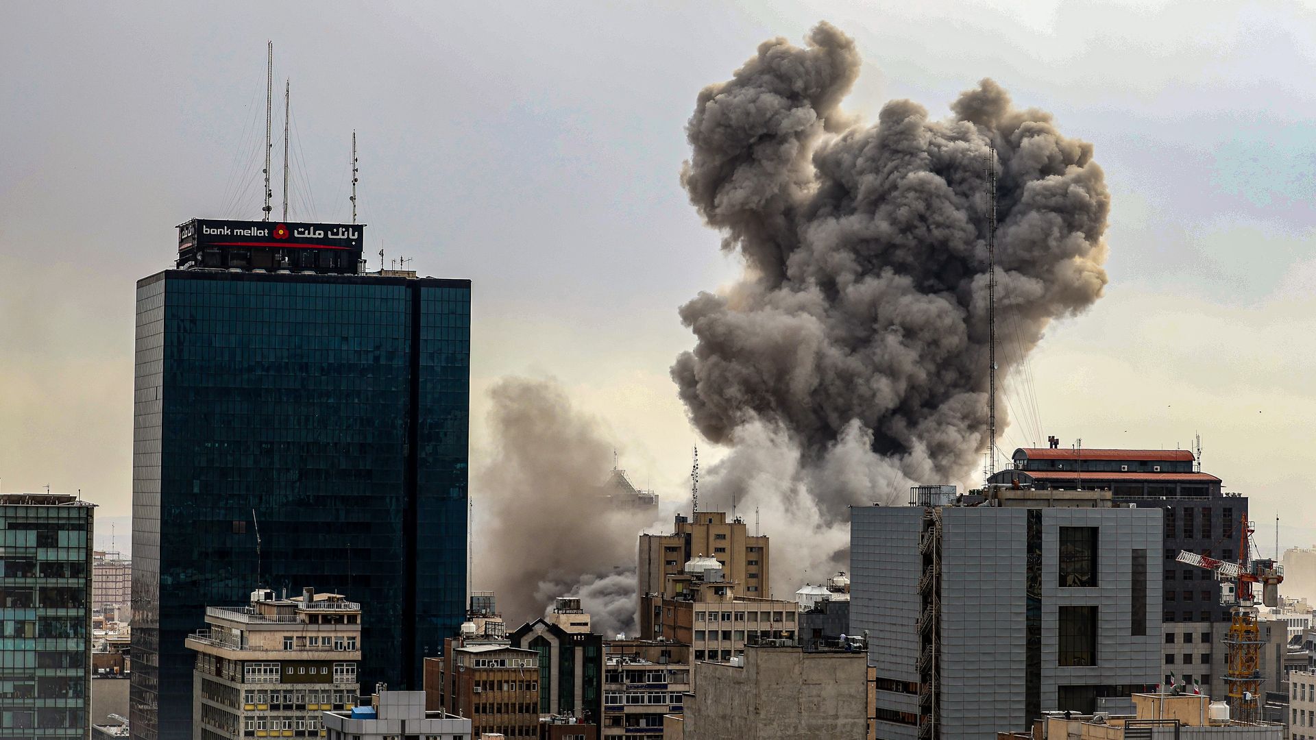 Distant view of Tehran cityscape with plumes of dark smoke rising above buildings after reported explosions.