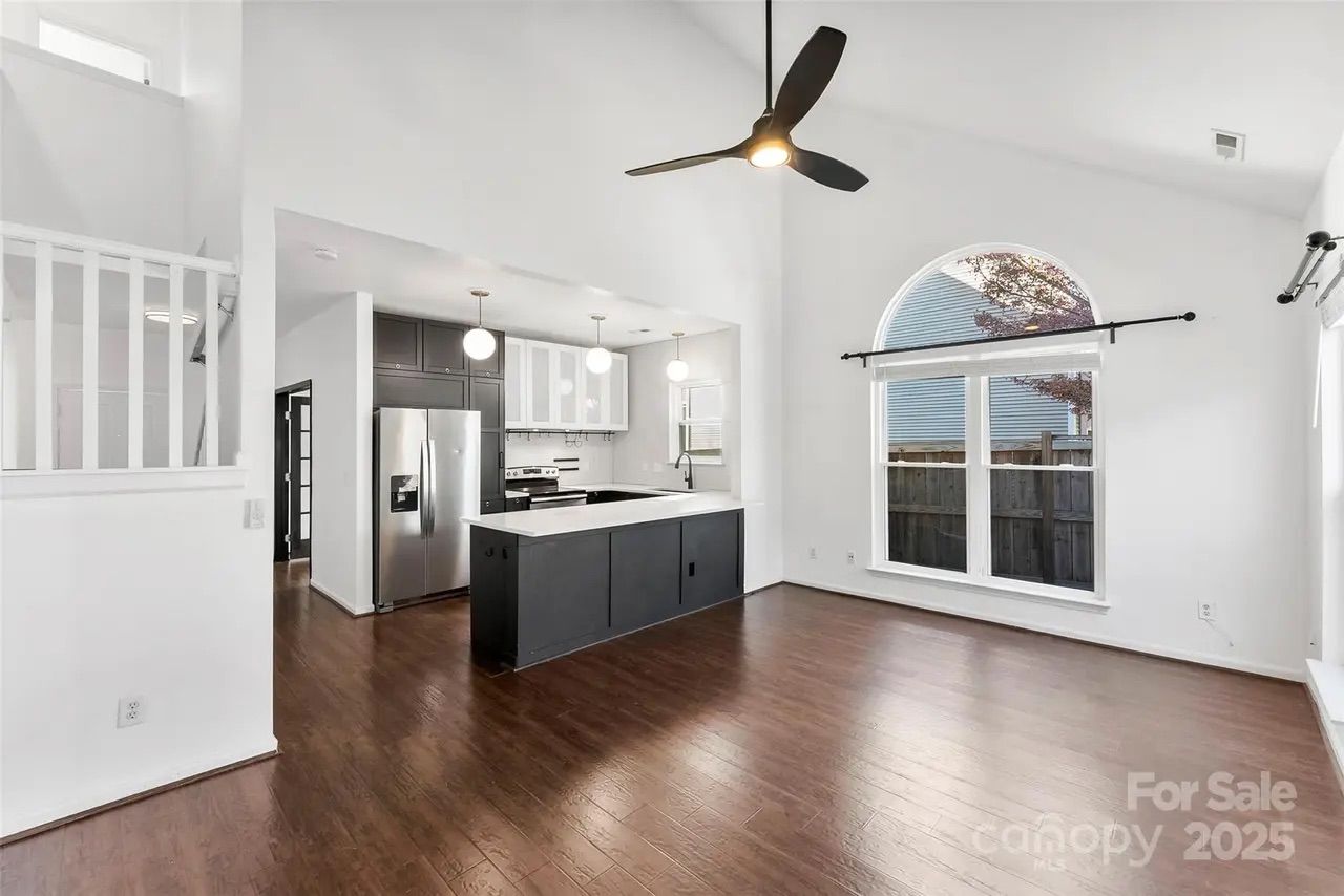 Bright living area with dark wood floors, white walls, a black ceiling fan, large arched window, and kitchen with black and white cabinetry and stainless steel appliances.