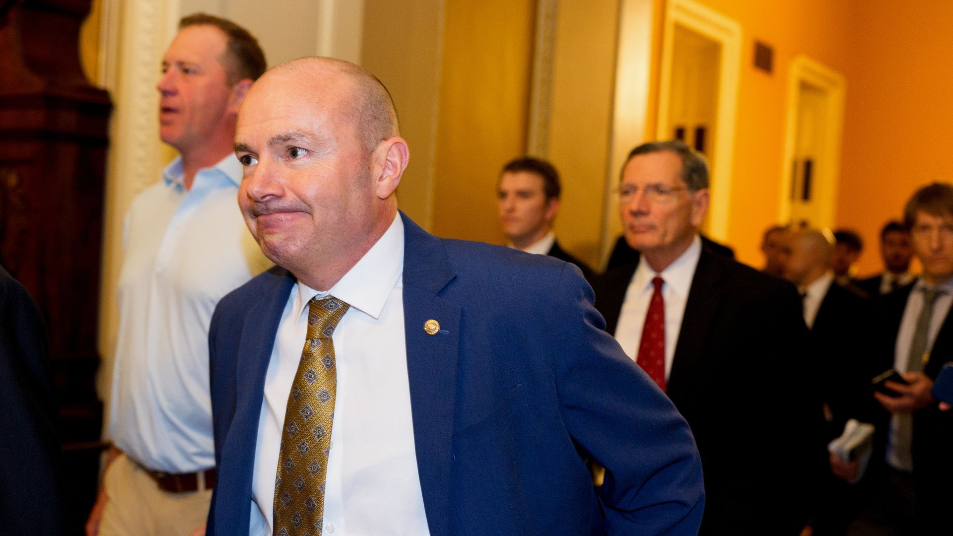 Group of men in suits and formal attire walking through a hallway with yellow walls; man in front wears blue jacket, white shirt, and patterned gold tie with a neutral expression.