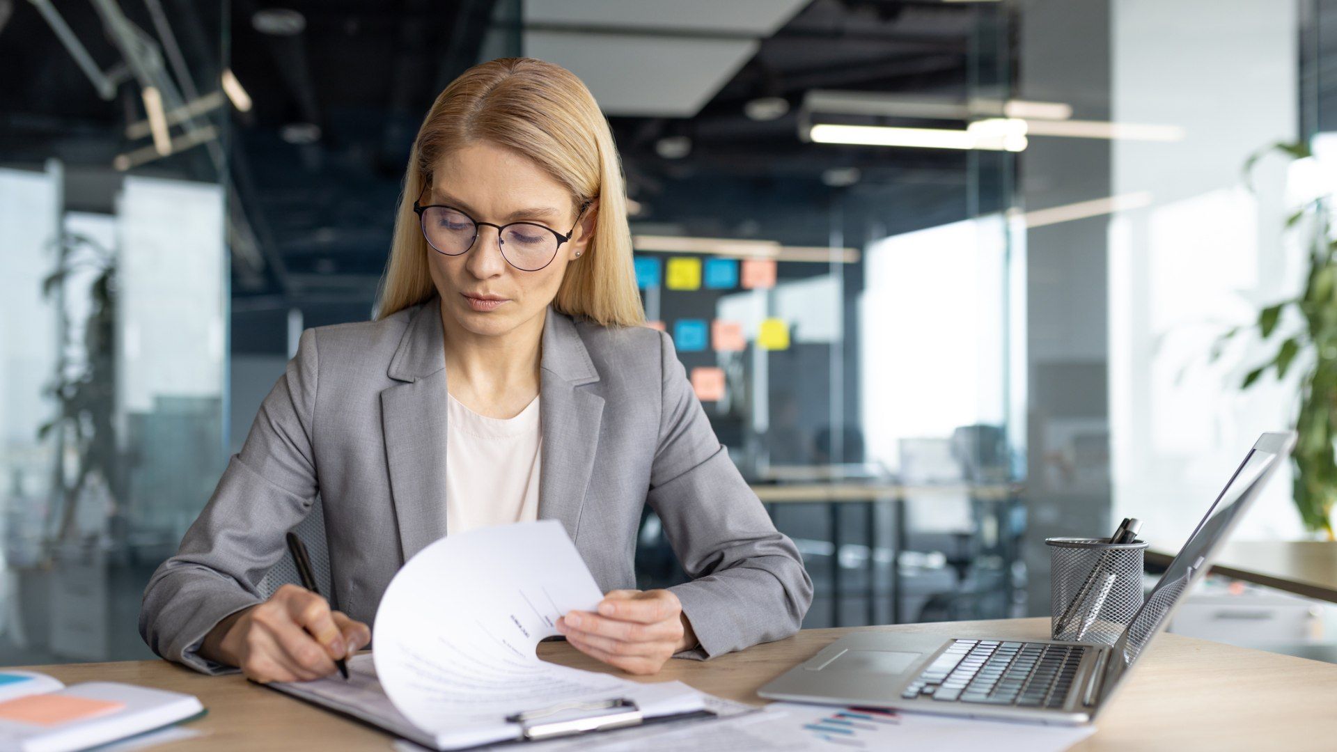 Blonde woman in a gray blazer and round glasses sits at a desk in a modern office, reading a document with a pen; a laptop and pencil cup sit nearby, with glass walls and colorful sticky notes in the background.