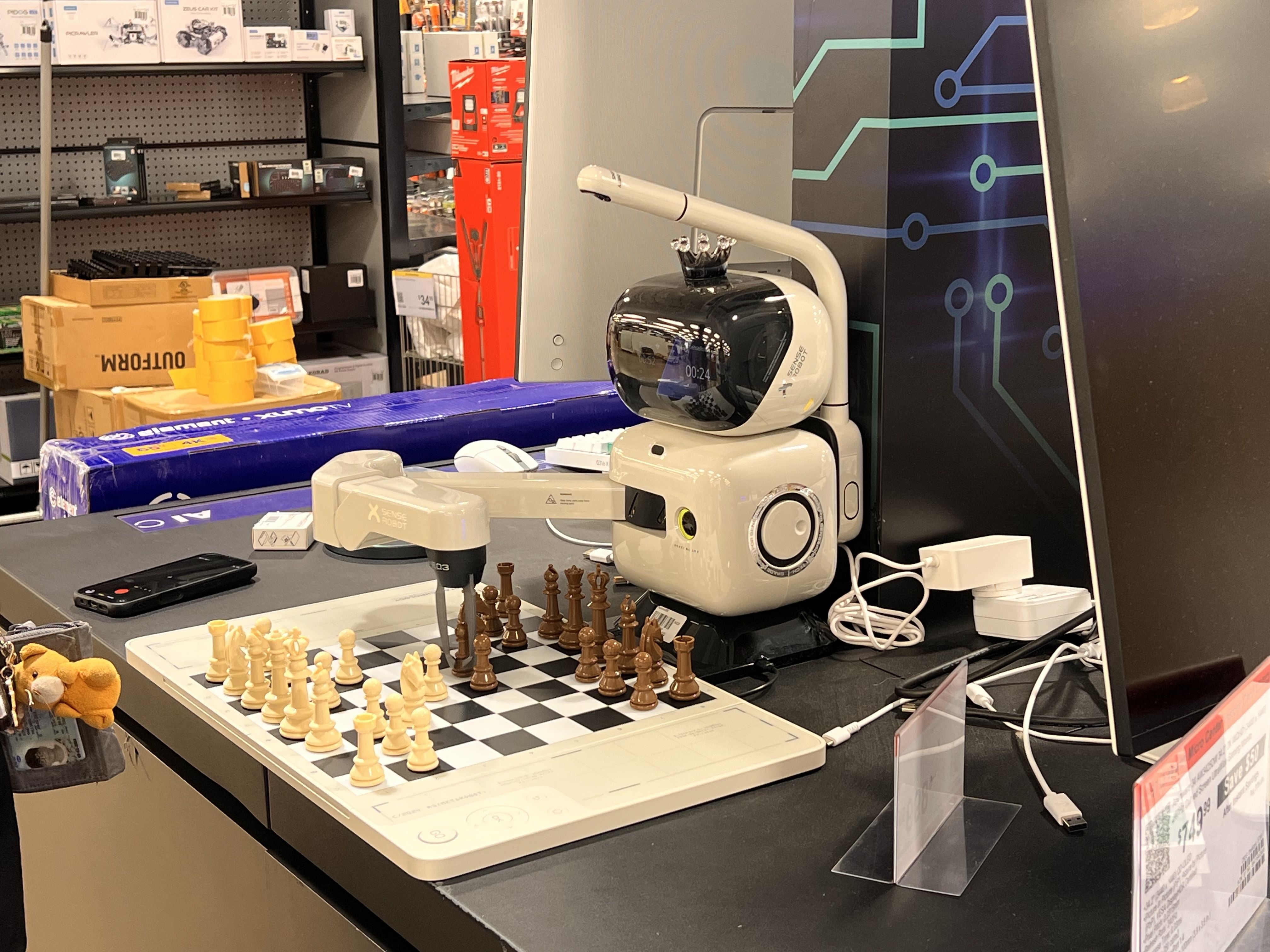 Store checkout area with a white curved-arm robot at the counter, a chessboard set with white and brown pieces, and shelves of boxed electronics in the background.