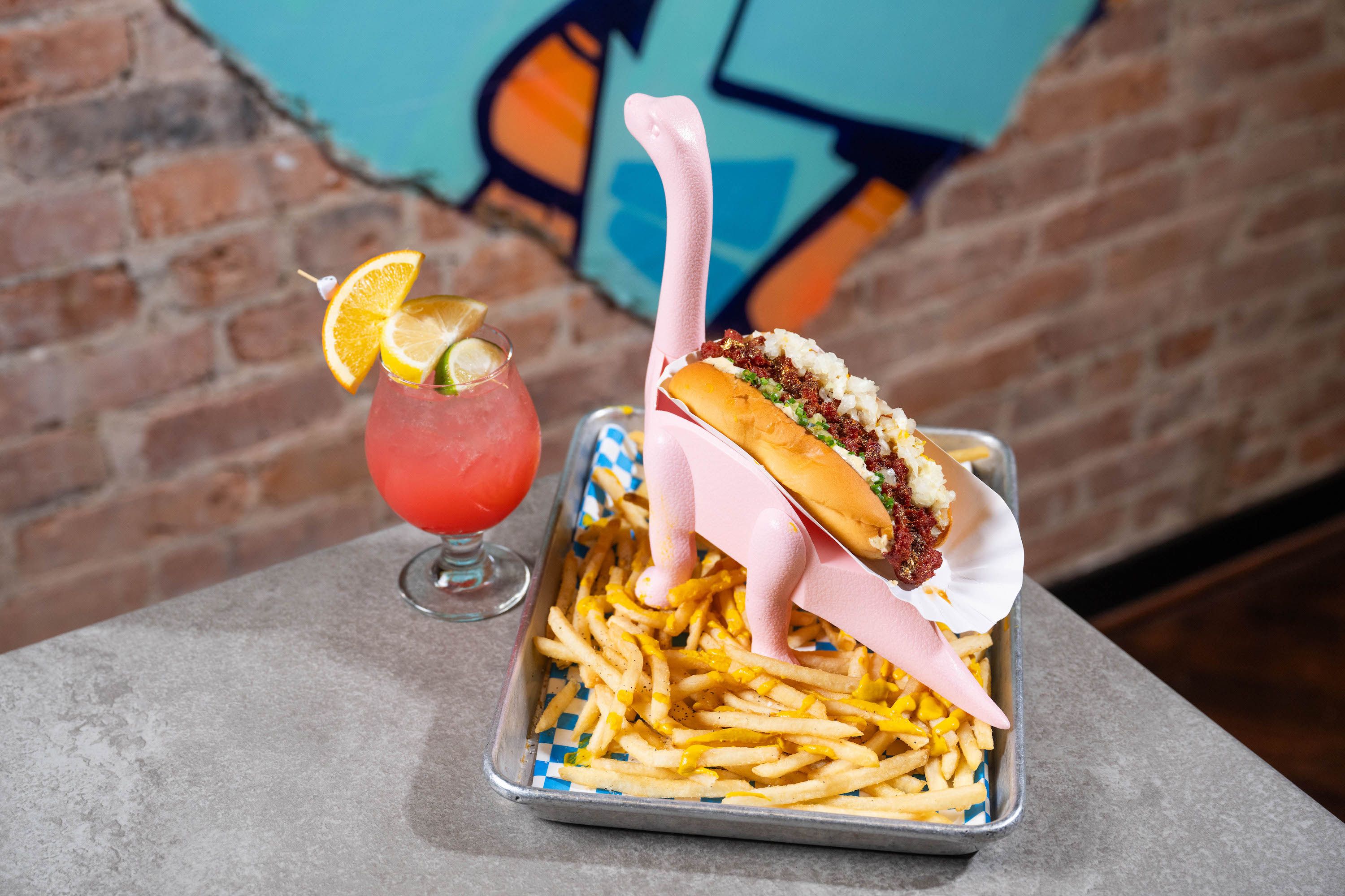 A pink dinosaur hot dog holder on top of French fries wit a cocktail on the side 