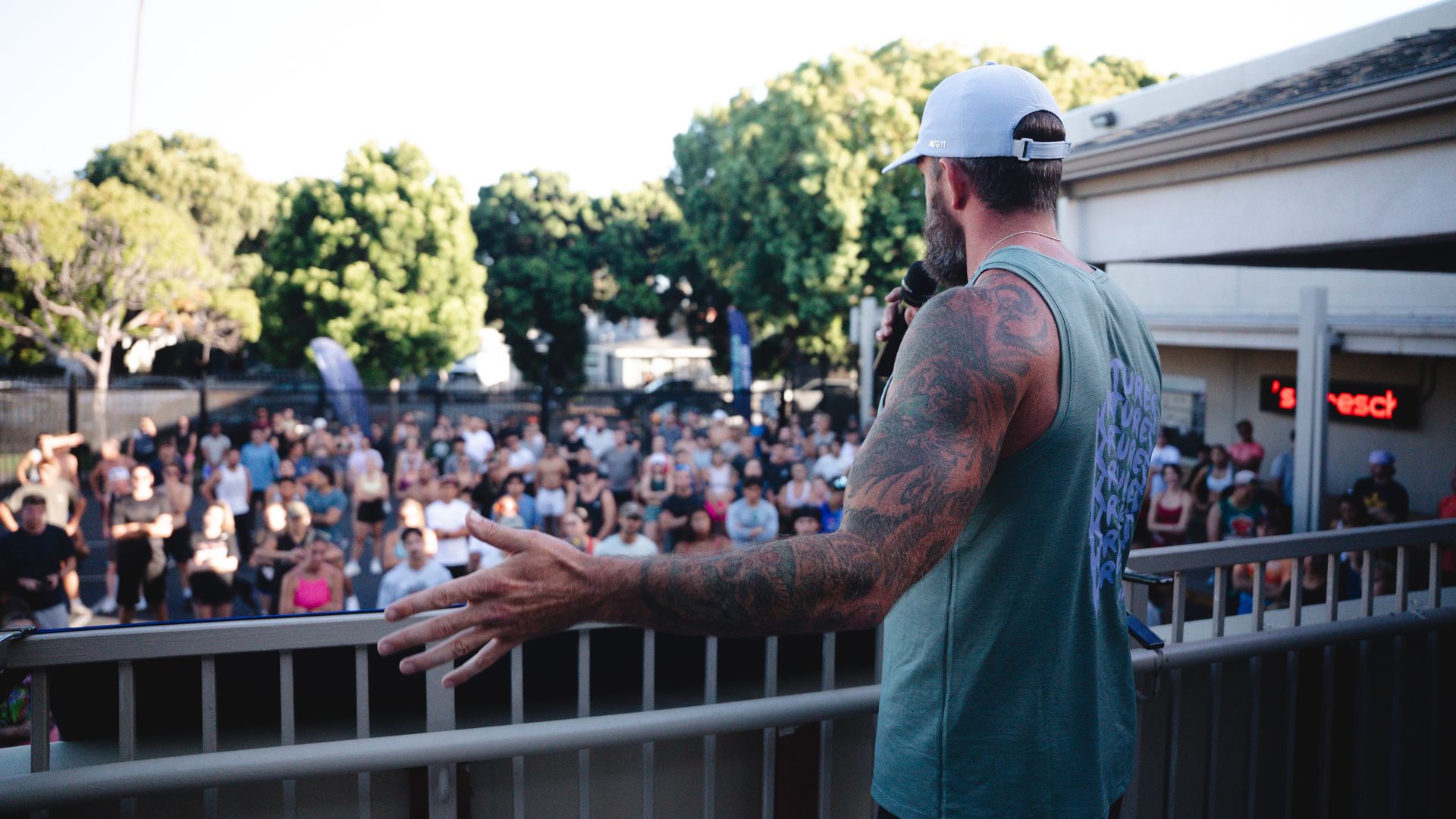 Side view of a tattooed man in a light blue cap and sleeveless teal shirt, holding a microphone and addressing a large outdoor crowd from a railing, with trees and a building in the background.
