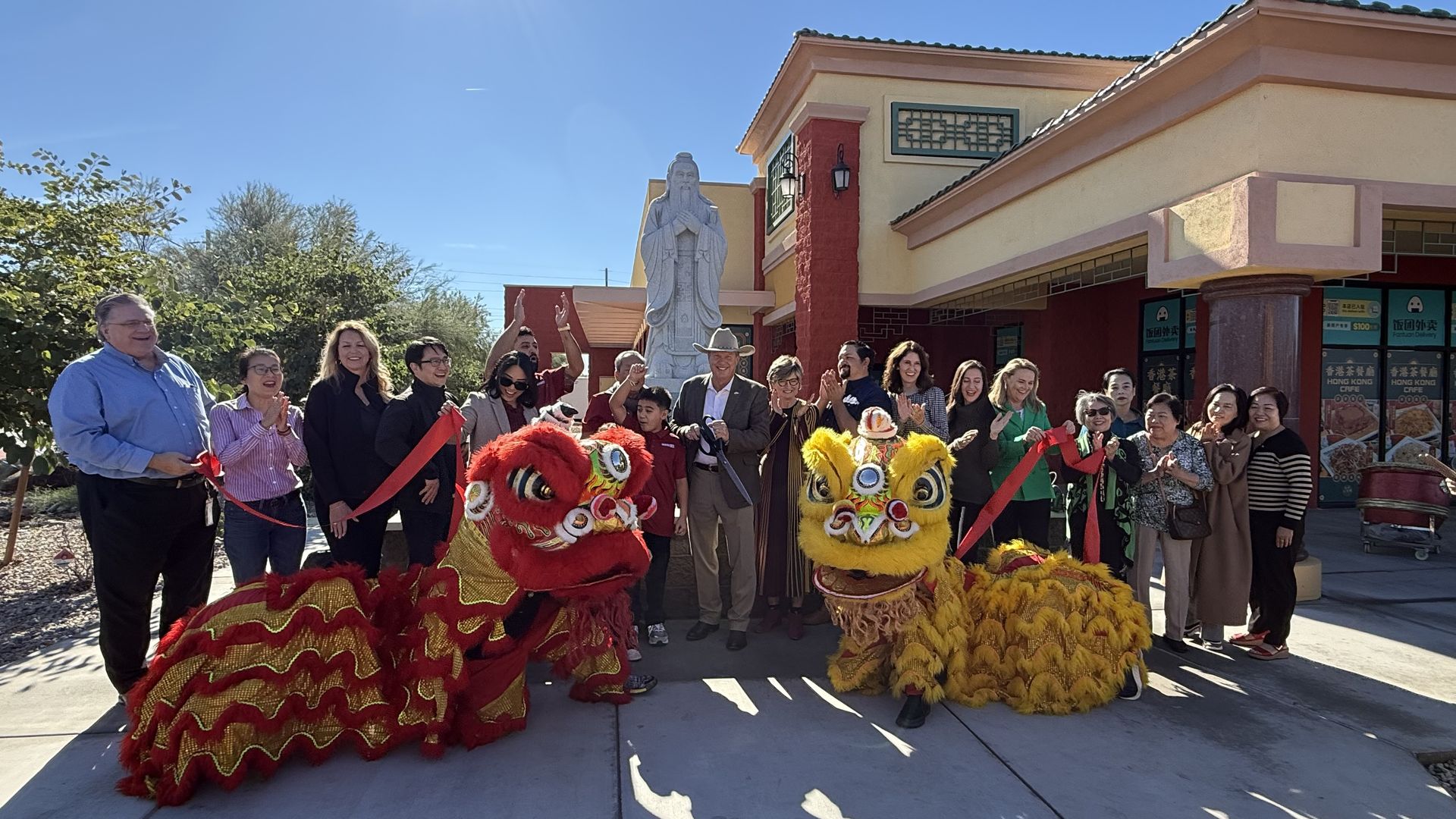 Group of people celebrating outdoors after cutting a large red ribbon, with two lion dance costumes, one red and one yellow, in front of a building and a large stone statue of Confucius next to a building. 