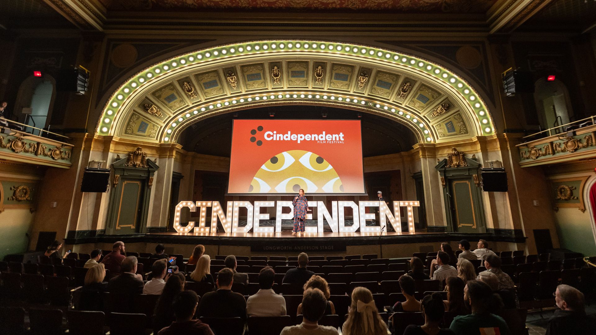An ornate theater with gold trim and a glowing arch of lights. A red screen reads "Cindependent Film Festival" as a performer stands on stage before a seated audience.
