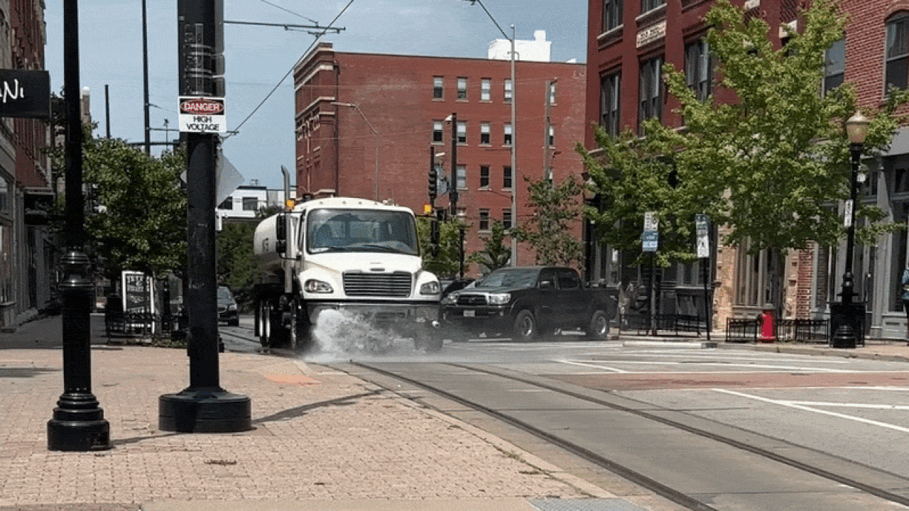 A white truck sprays water at an urban intersection, creating a spray behind it. A black SUV is parked nearby; brick buildings, trees, and overhead tram lines frame the street.