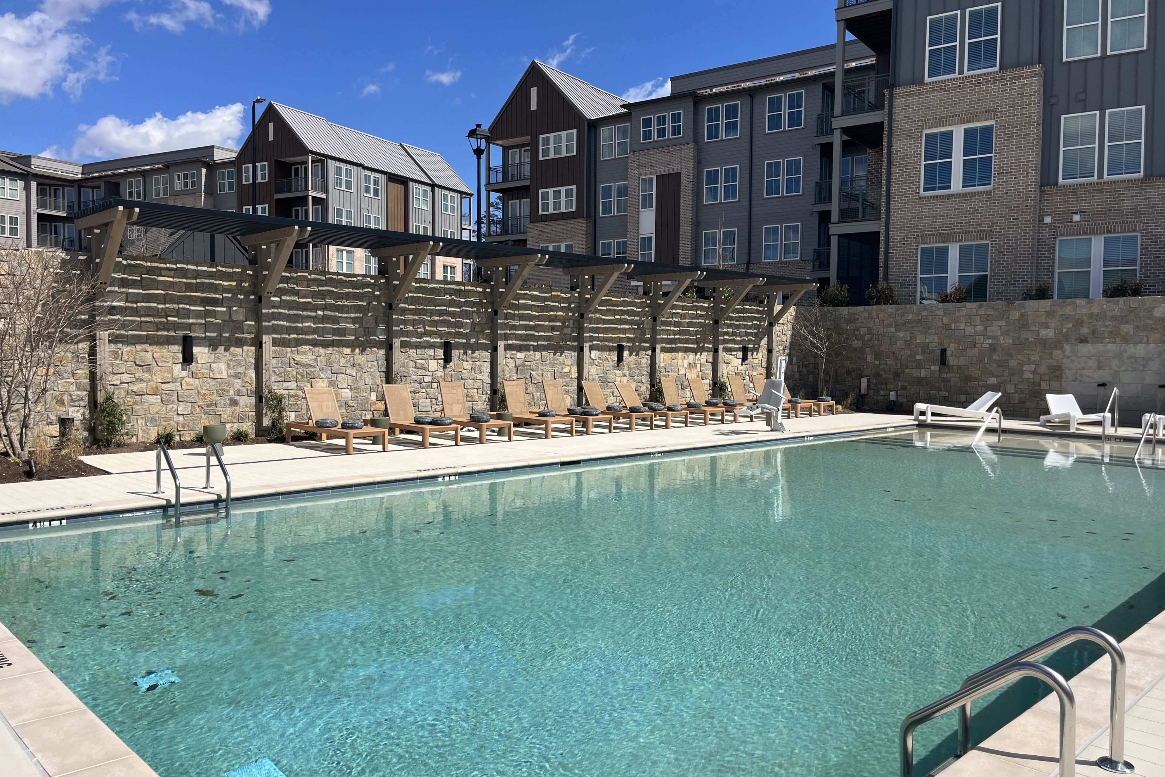 Pool area at a modern apartment complex: clear blue water, tan lounge chairs along a stone wall under a pergola, with multi-story gray-brick buildings in the background.