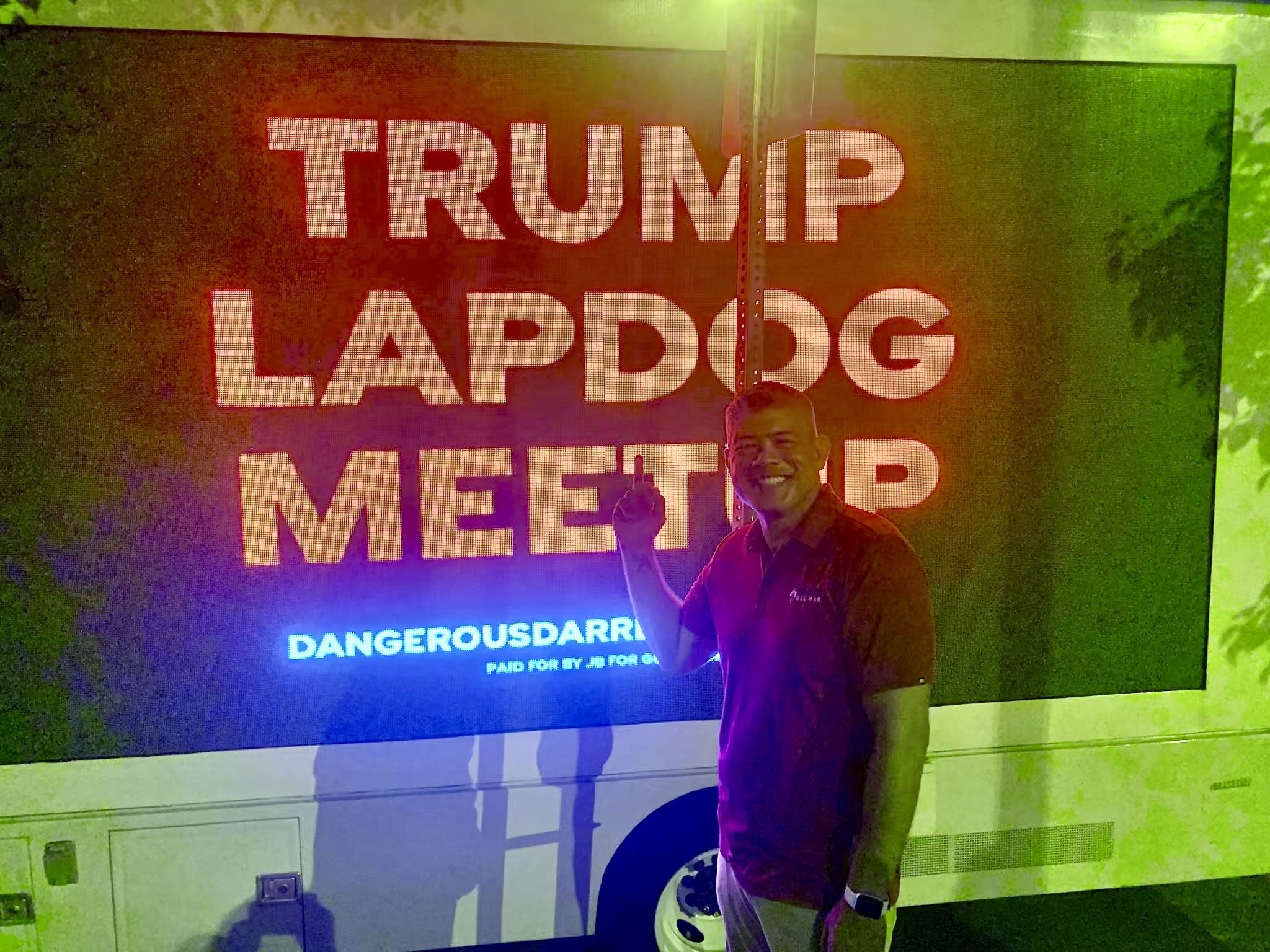 Smiling man in a red shirt poses a selfie in front of a large screen that says "TRUMP LAPDOG"; bright dotted letters and vivid green, red, and blue lighting.