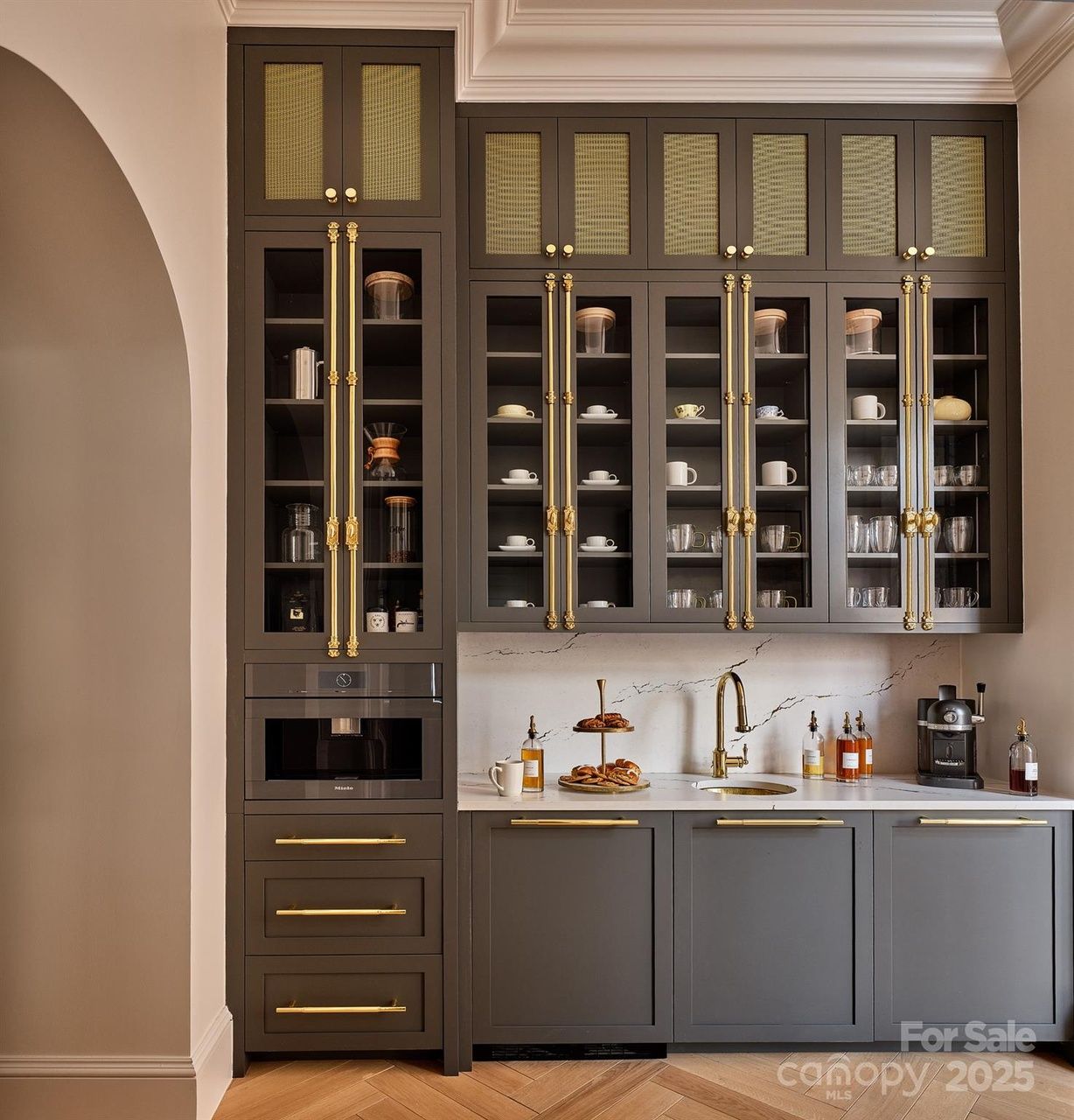 Modern kitchen with matte gray cabinets, gold handles, glass doors, marble backsplash, gold faucet, pastries on a tiered tray, and countertop coffee setup with machine and syrups.