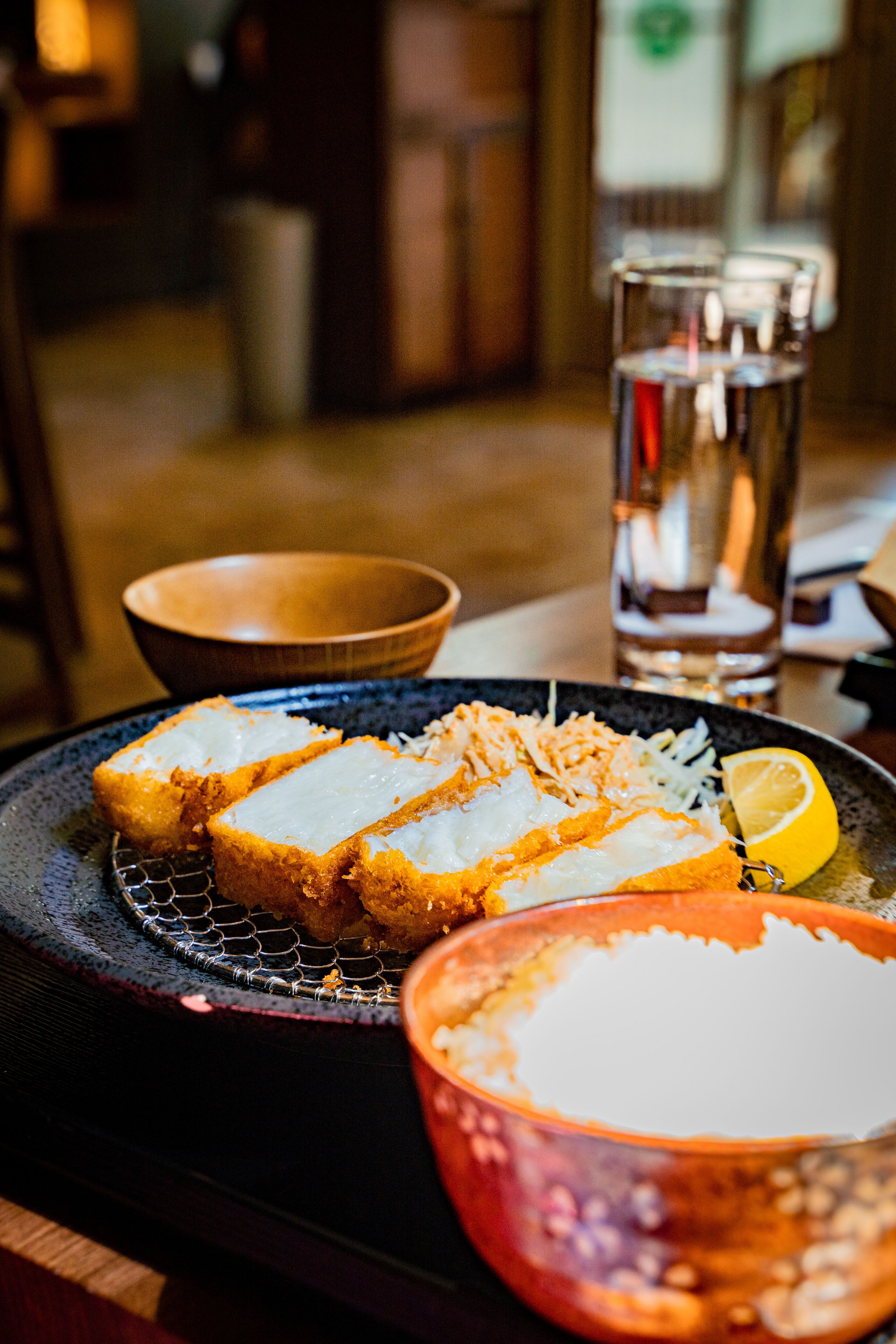 Breaded fried fish slices on a dark plate with a lemon wedge, atop a wire rack with shredded cabbage; a glass of water and warm blurred restaurant interior in the background.