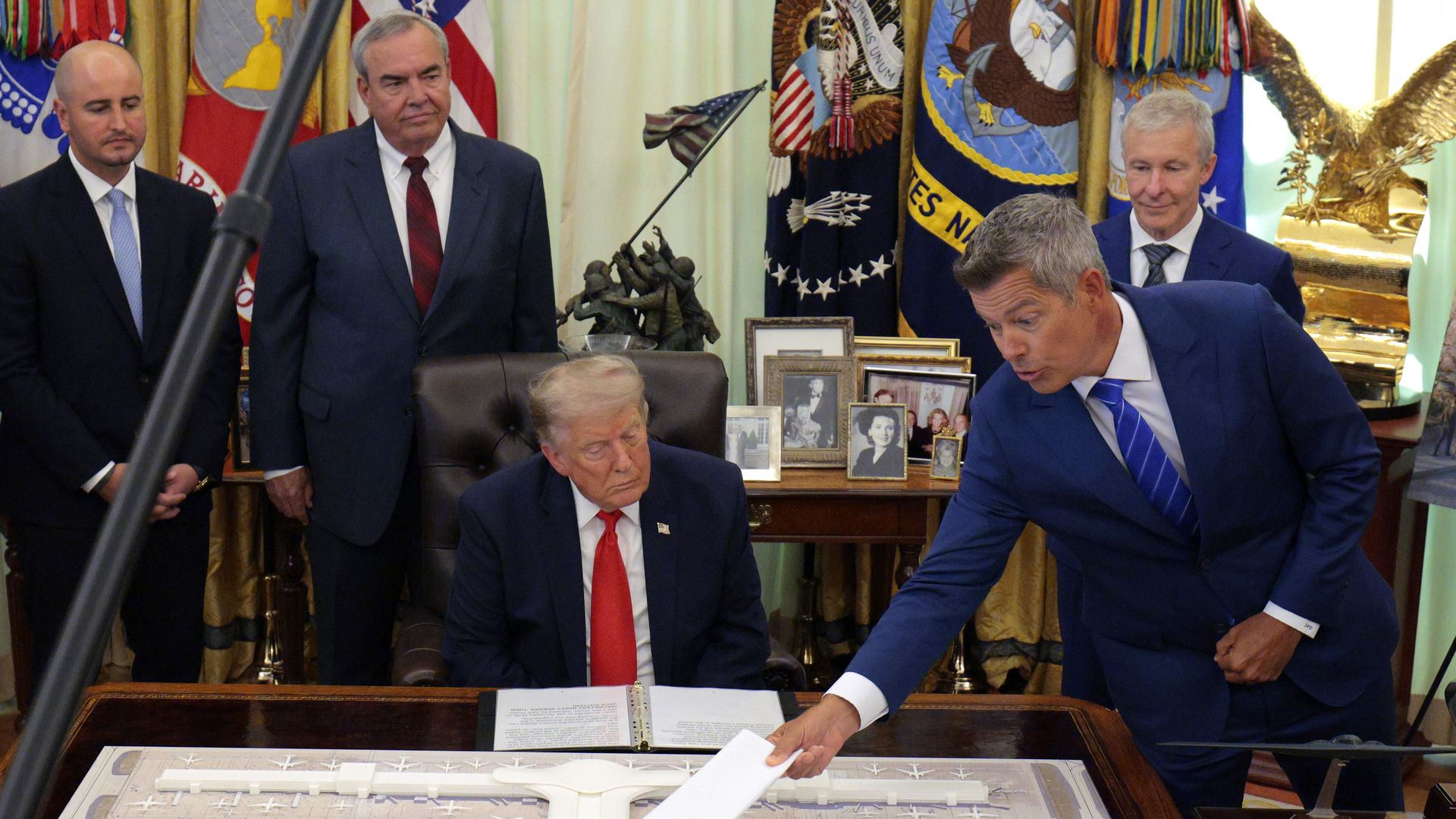 An image of President Trump holding a laser pointer over a physical mockup of the renovated Dulles Airport, with Jack Potter and Transportation Secretary, among others, standing behind him