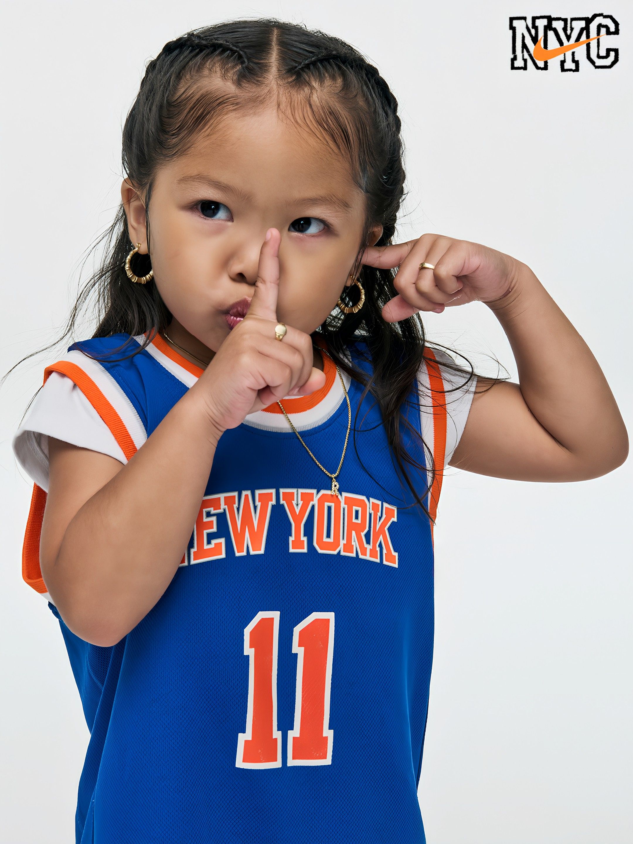 Young girl in a blue New York basketball jersey with the number 11, making a shushing gesture with her finger to her lips; braided hair, gold hoops, NYC logo in the corner.