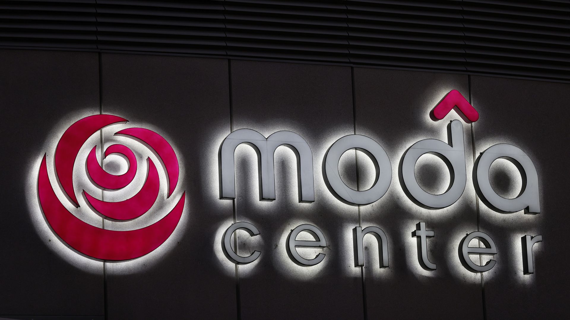 Exterior of the Moda Center at nightt: a large pink magenta circular logo with white inner swirl on the left, and gray 3D letters spelling "Moda Center" along a dark wall.