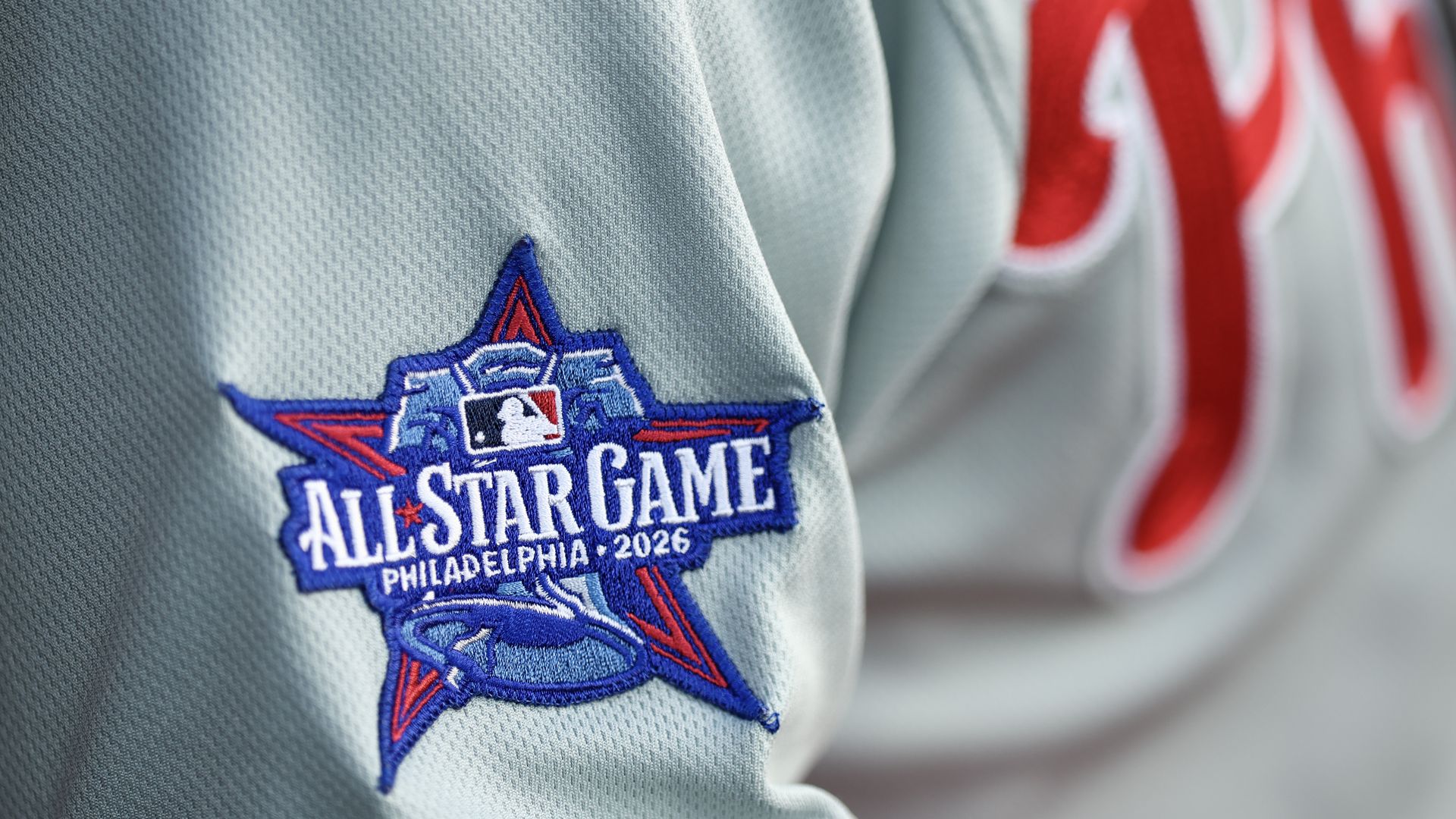 Close-up of light-gray, textured jersey fabric with a blue embroidered star patch that reads "ALL-STAR GAME" and "PHILADELPHIA 2026"; red lettering blurs in the background.