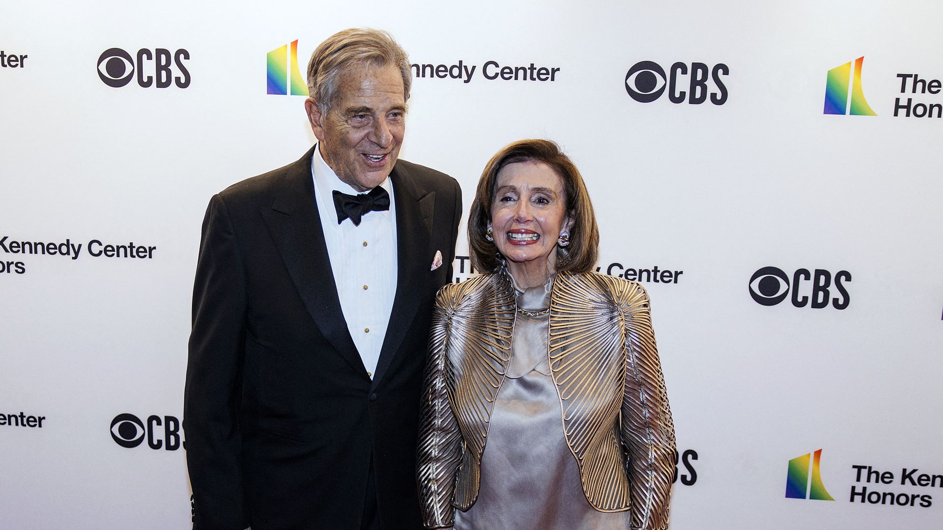House Speaker Nancy Pelosi wearing a gold outfit and her husband Paul Pelosi wearing a Tuxedo in front of a wall bearing the CBS and Kennedy Center logos.