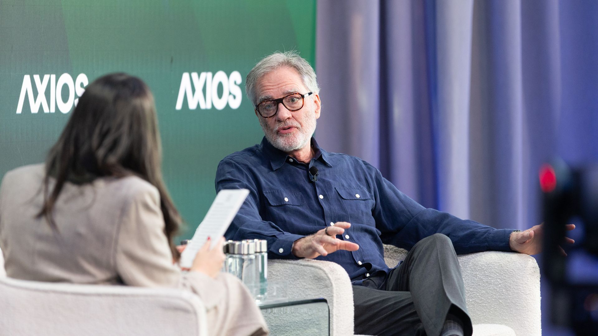 An older man with glasses and a gray beard in a navy shirt speaks during an Axios interview, seated in a light gray chair; a woman interviewer with a notepad faces him. Green Axios backdrop.