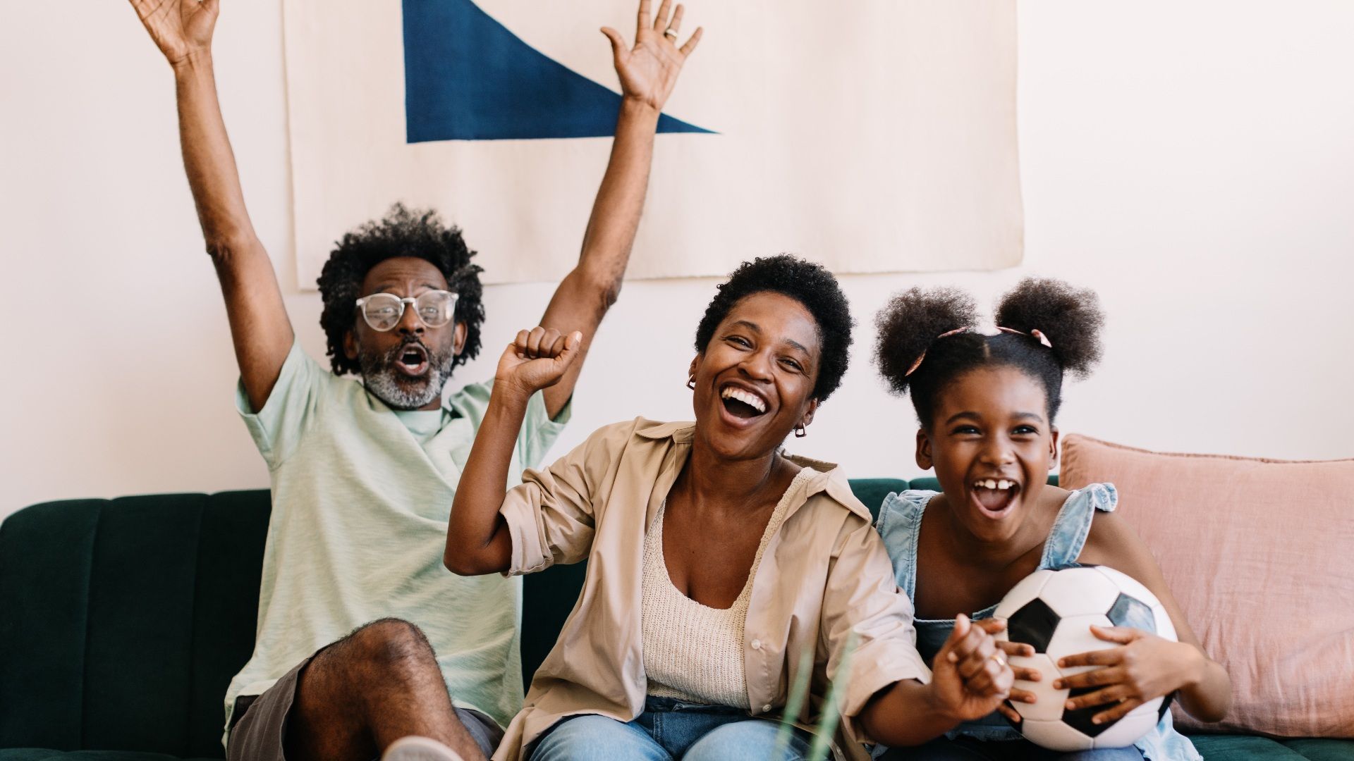 Cheering family on a living room sofa: an older man with glasses raises his arms, a smiling woman, and a girl with a soccer ball, as a blue banner hangs on the wall behind them.