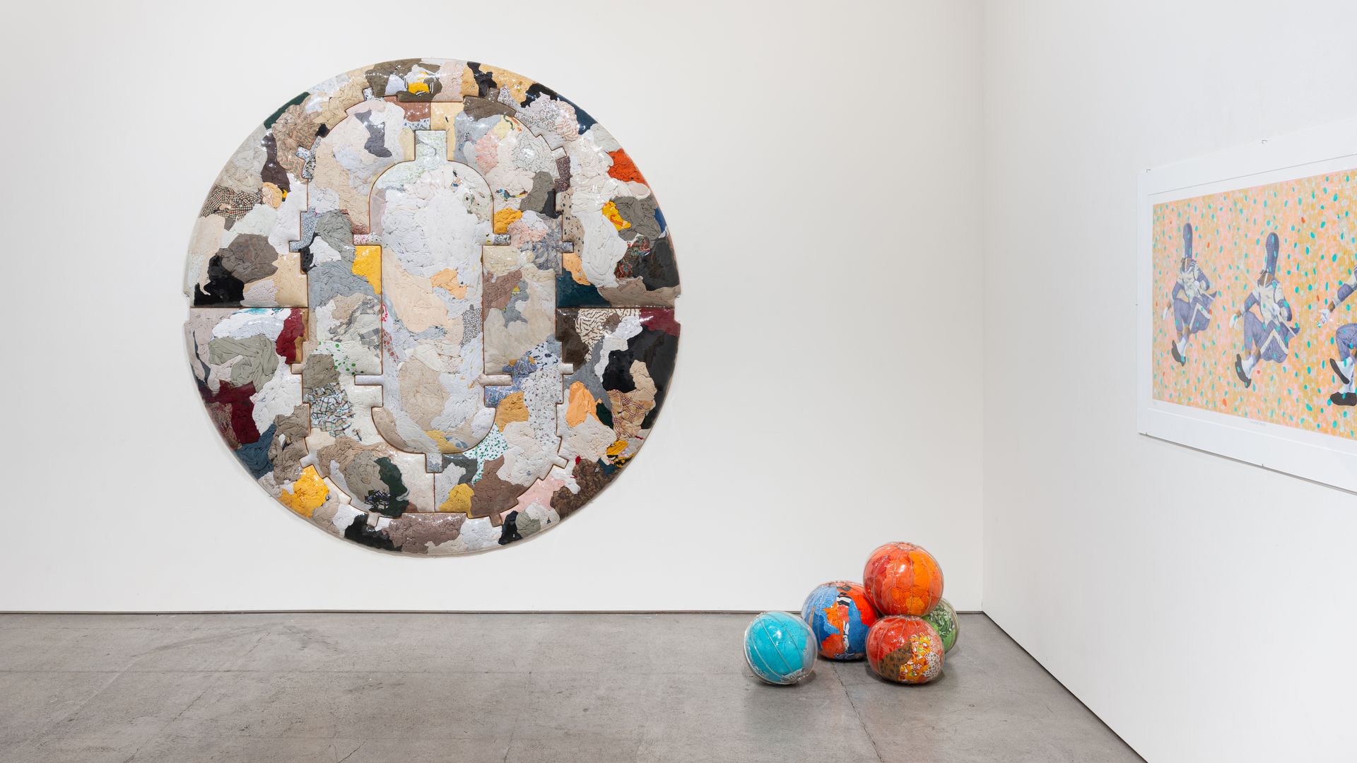 White gallery with a large circular mosaic made of irregular colored patches on the left, a framed orange-and-blue patterned artwork on the right, and several painted globes on the floor.