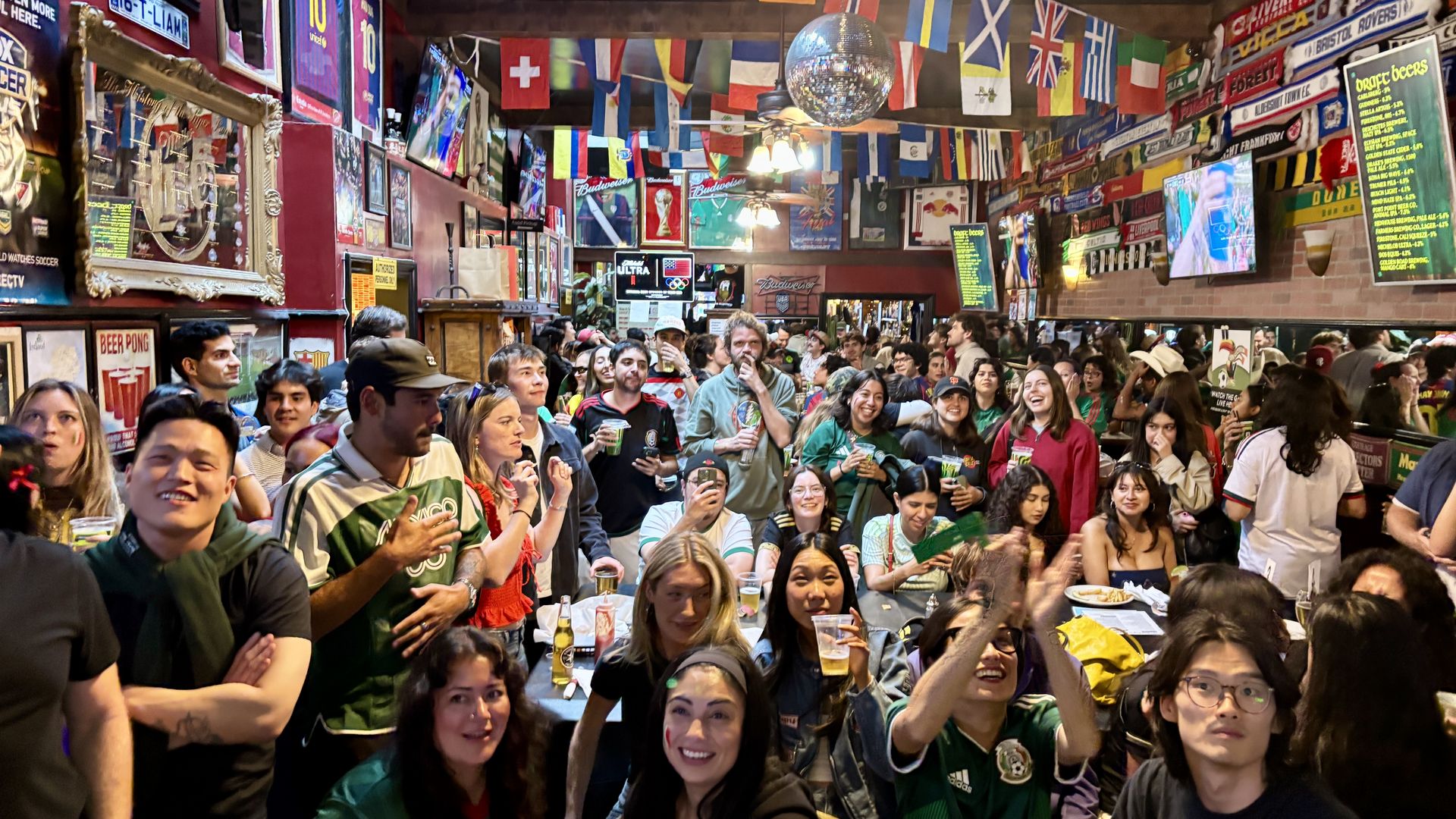 Busy sports bar filled with a large crowd holding drinks, many wearing green jerseys. Colorful flags hang from the ceiling, a disco ball overhead, and multiple screens show sports.
