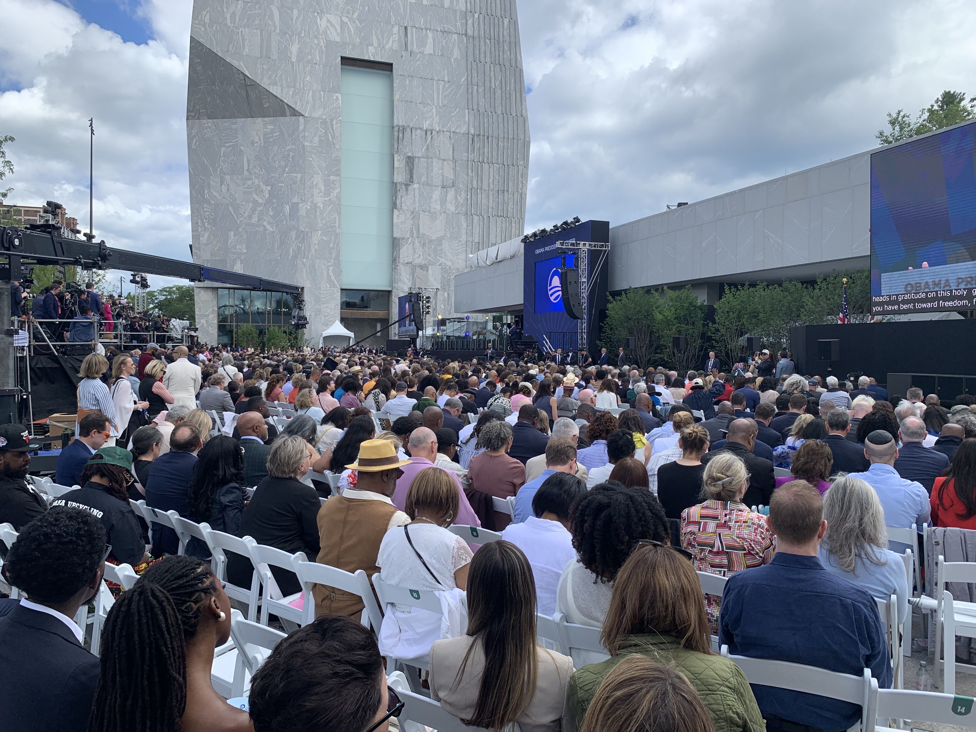 Large outdoor ceremony with a crowded audience facing a stage and a large LED screen; a camera crane and media on the left; tall marble building behind; cloudy sky; screen shows Obama signage.