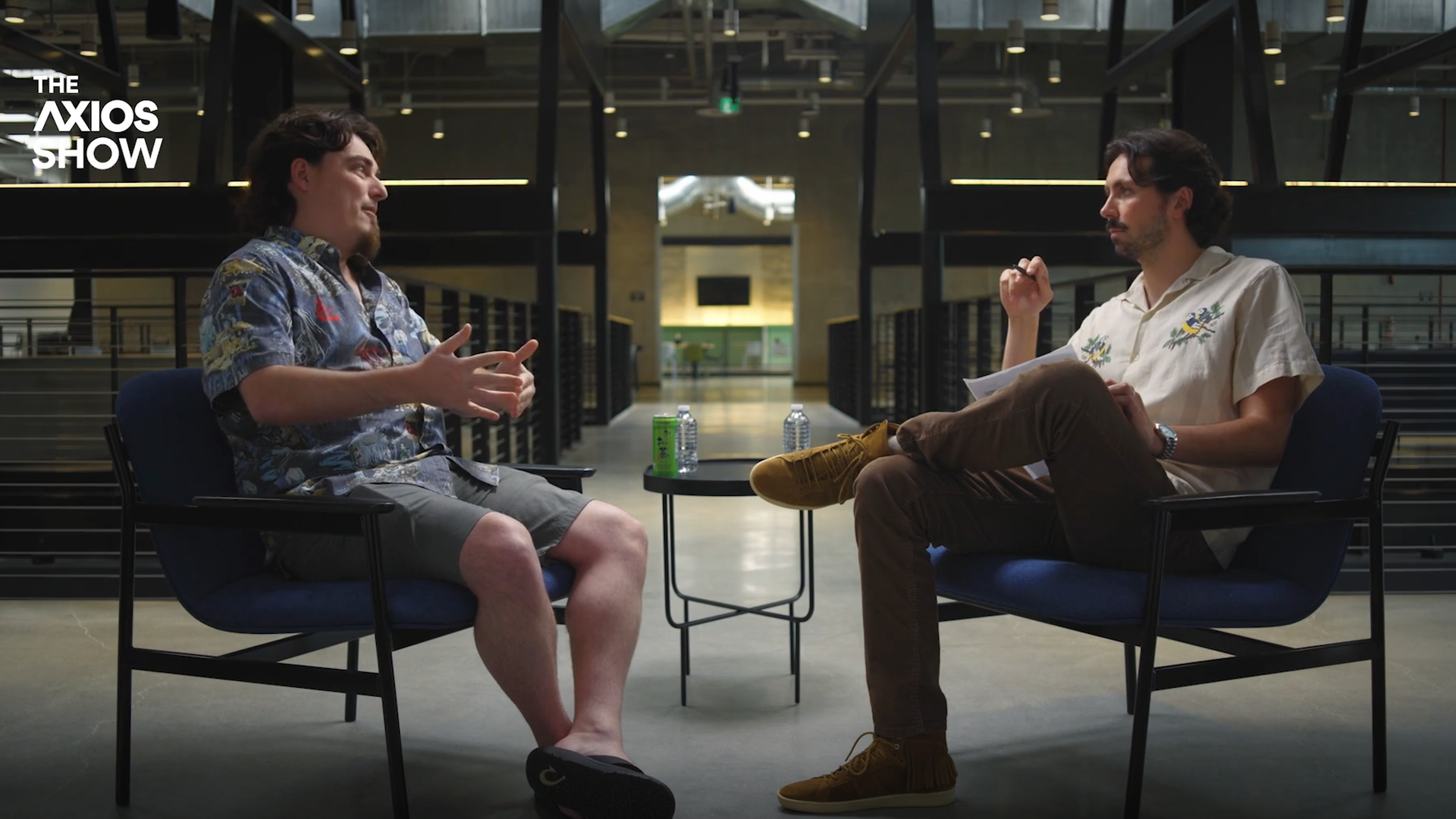 Palmer Luckey and Colin Demarest sit in blue chairs facing each other in a modern interior; one in a tropical shirt and shorts, the other in a light floral shirt with papers.