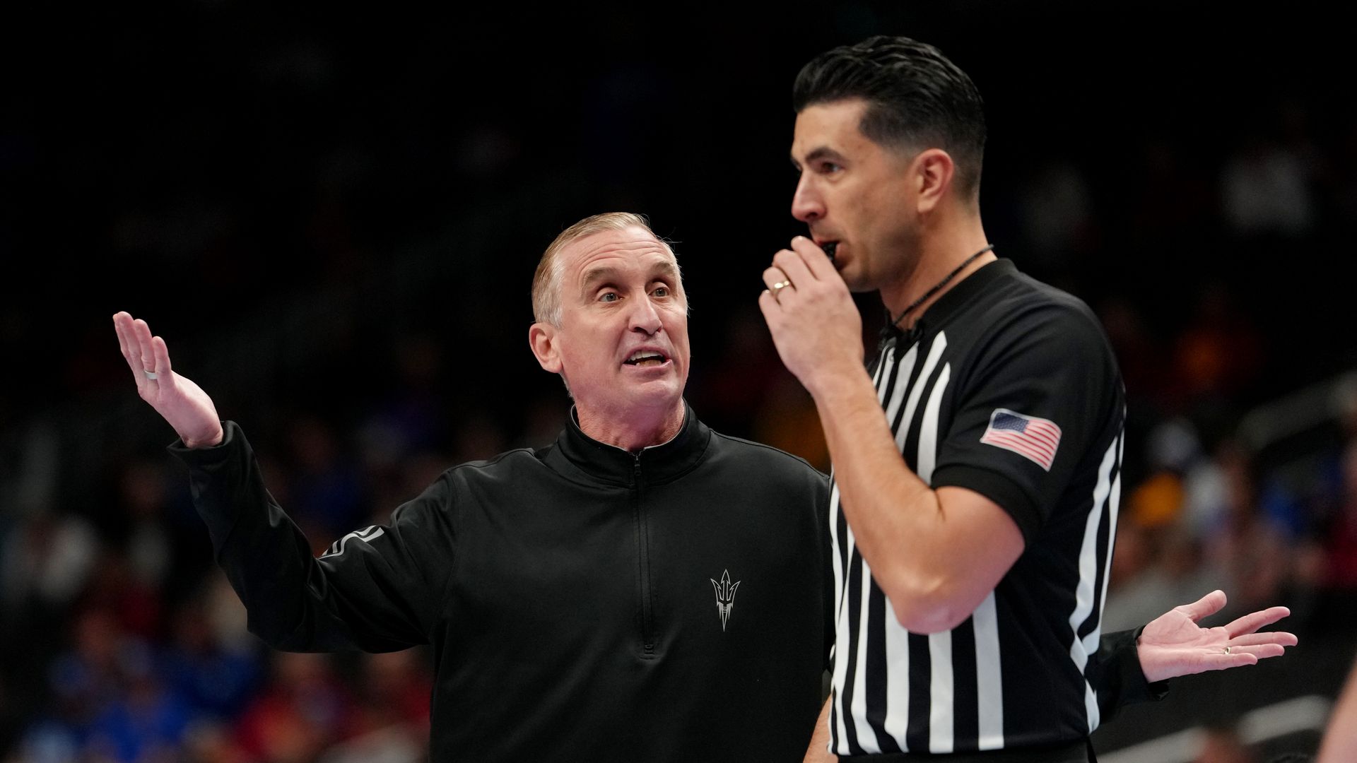 A man in a black zip-up with a pitchfork logo on his left side holds his hands to his side and argues with a referee in a black and white striped shirt and a whistle in his mouth. 