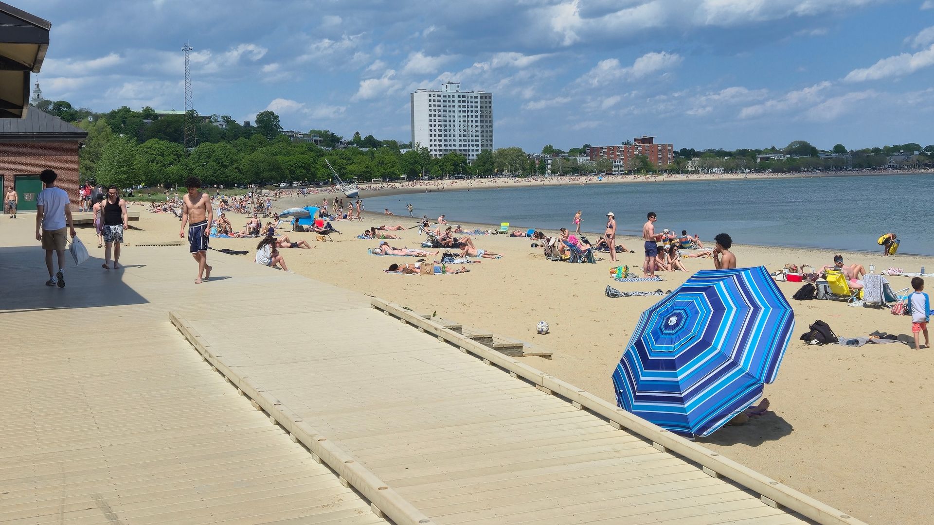 Busy city beach with sunbathers on sand, blue striped umbrella, and a wooden boardwalk in the foreground; skyline includes a tall white high-rise, green trees, and a calm bay.