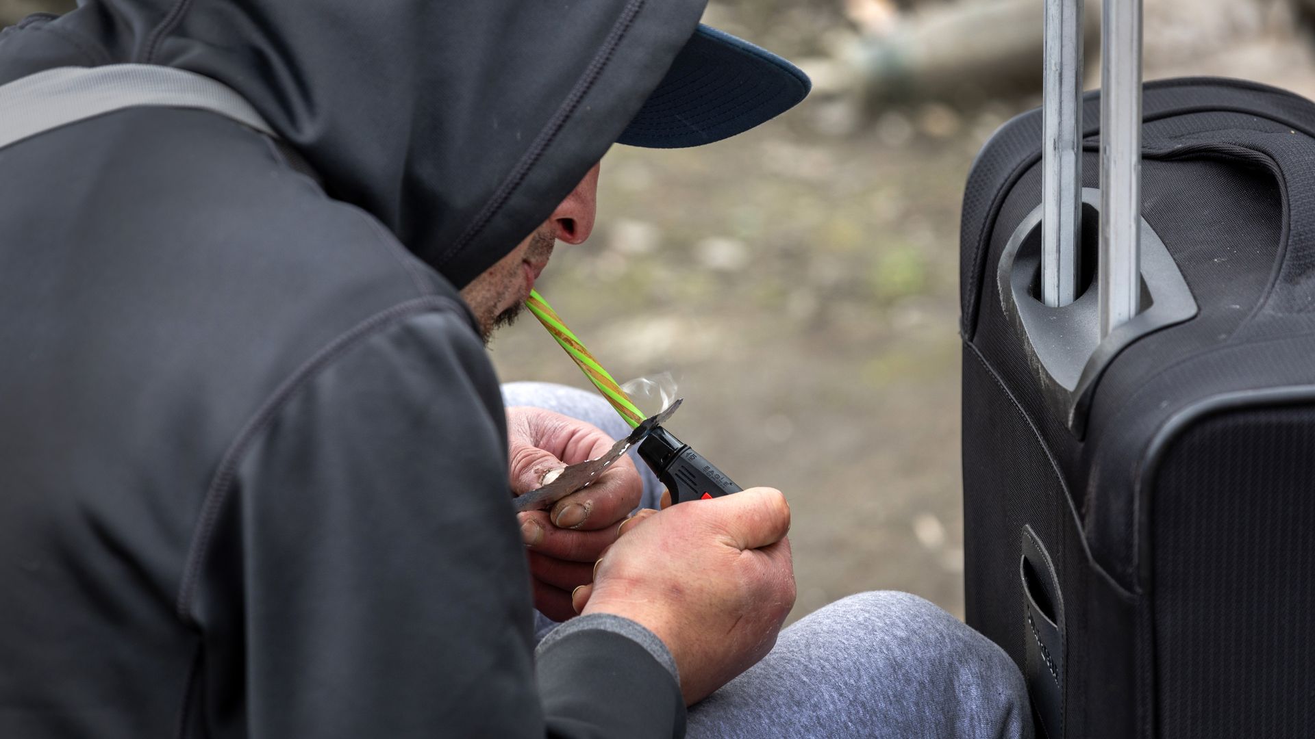 A photo of a man smoking fentanyl outside, sitting down next to a suitcase.