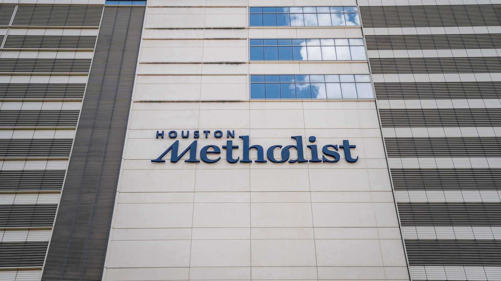 Exterior facade of a tall building featuring the blue signage "Houston Methodist" on a beige wall, with reflective glass windows and metal louvers under a partly cloudy sky.