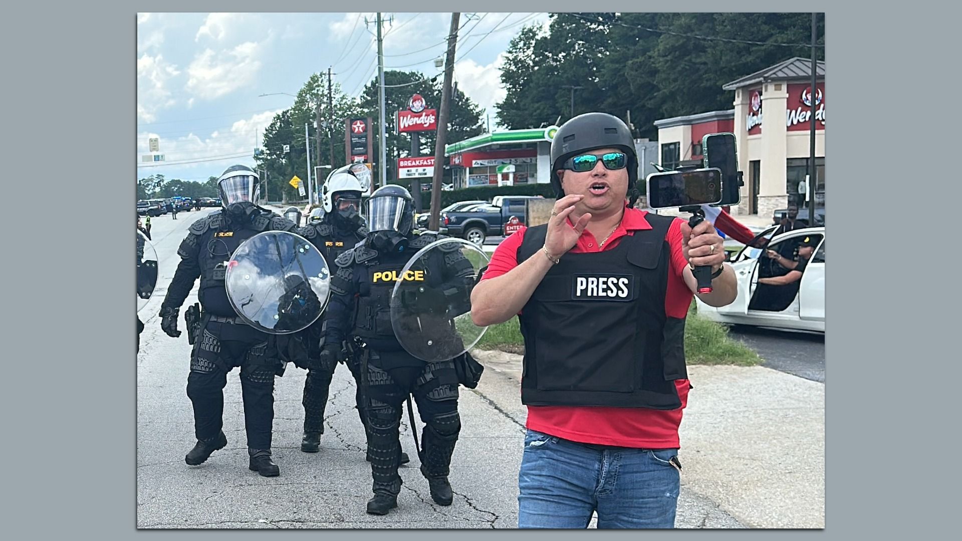 A man wearing a black press vest and helmet holds a phone on a stabilizer, speaking. Behind him, police in full riot gear with shields walk on a street near a Wendy's and gas station.