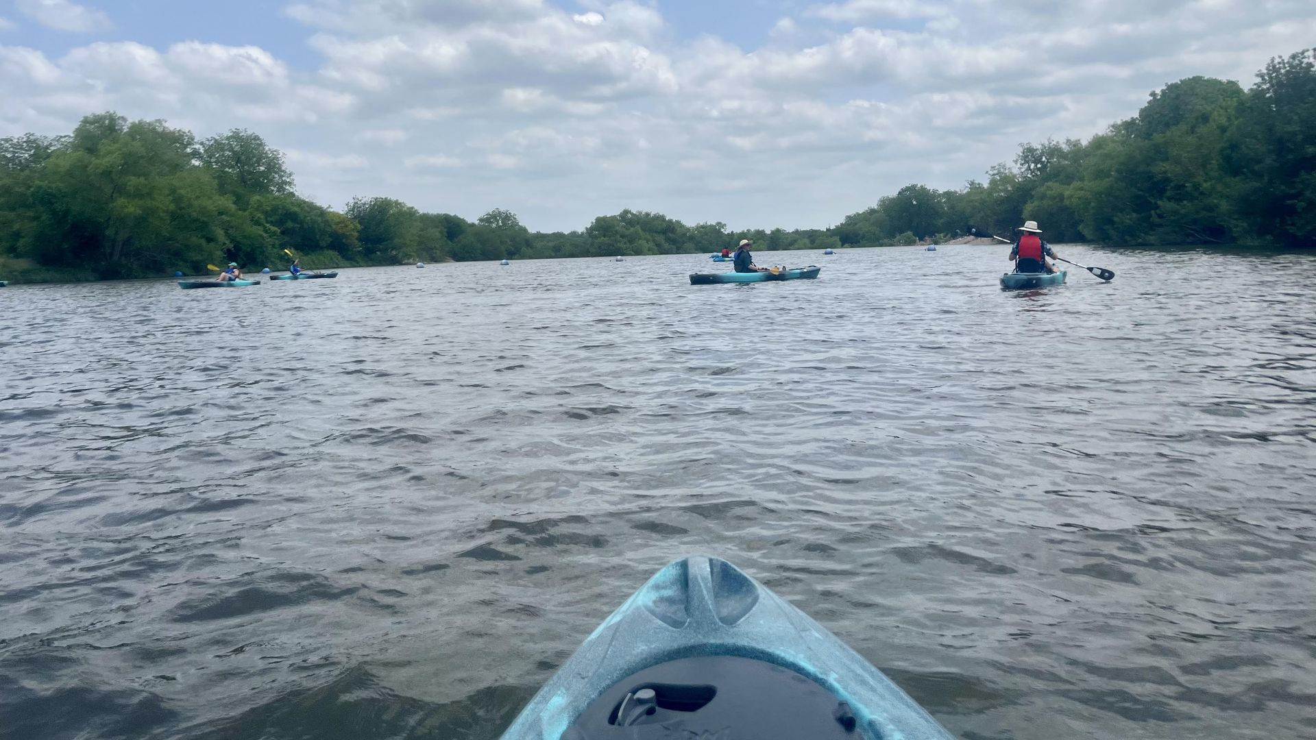 A view of the San Antonio River from within a kayak shows other kayakers in the distance near trees in the water.