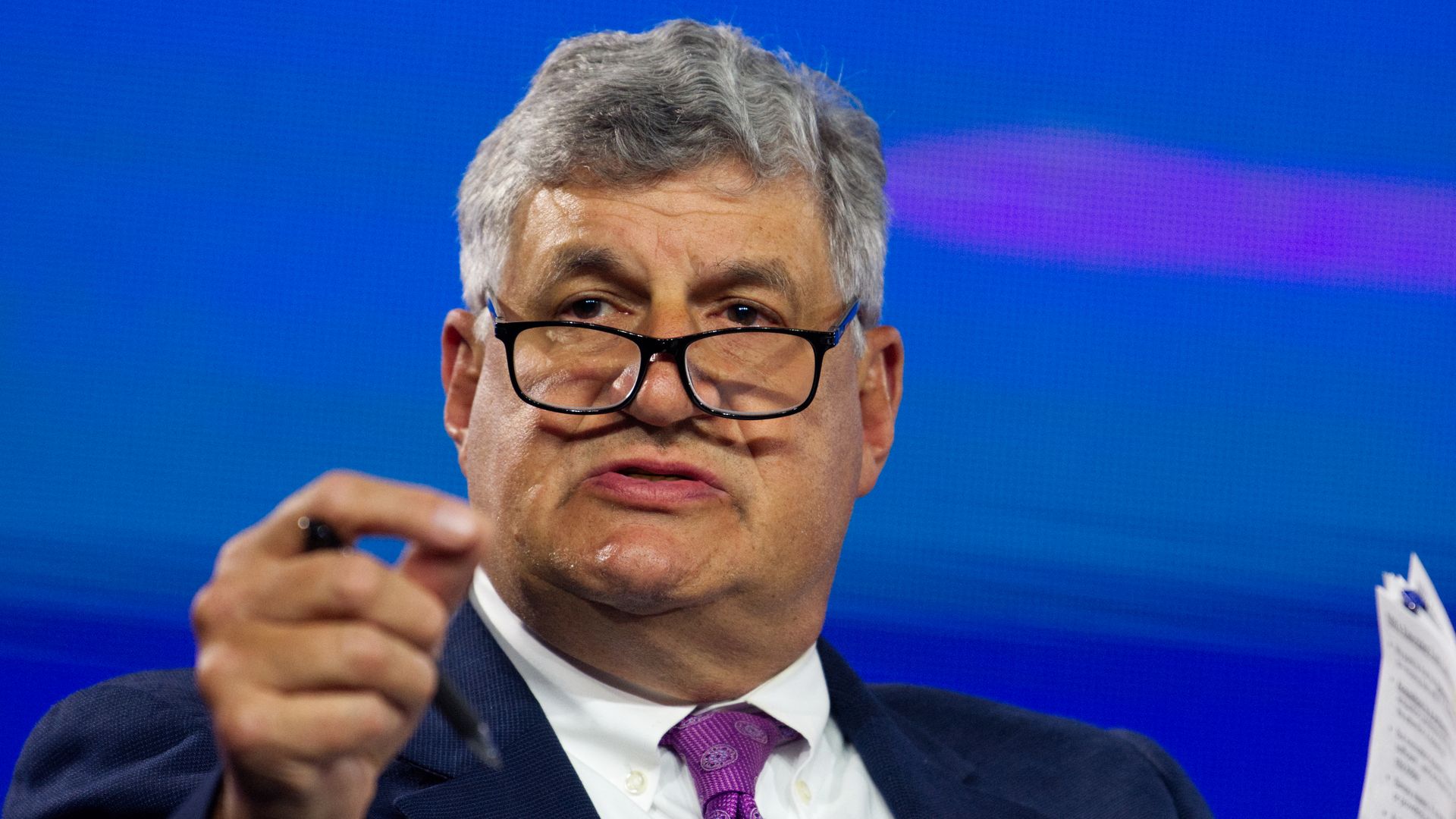 Bill LaPlante, a man, gestures and holds up his notes as he speaks at an event. He is in a dark suit, white shirt and wearing a purple tie.