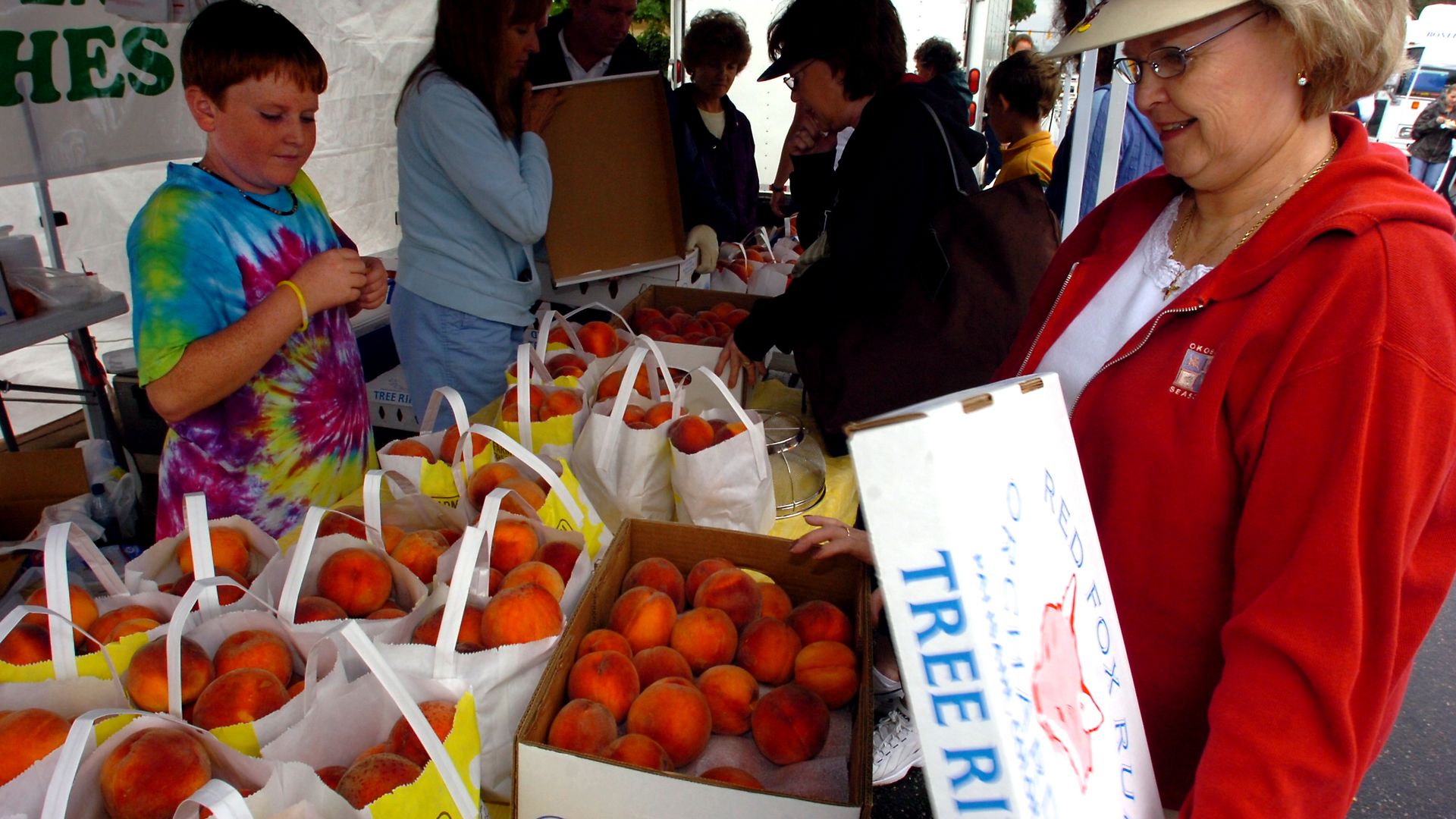 Outdoor market stall under a tent with baskets of peaches in white bags. A boy in tie-dye on the left and a smiling woman in red on the right, shoppers in the background.