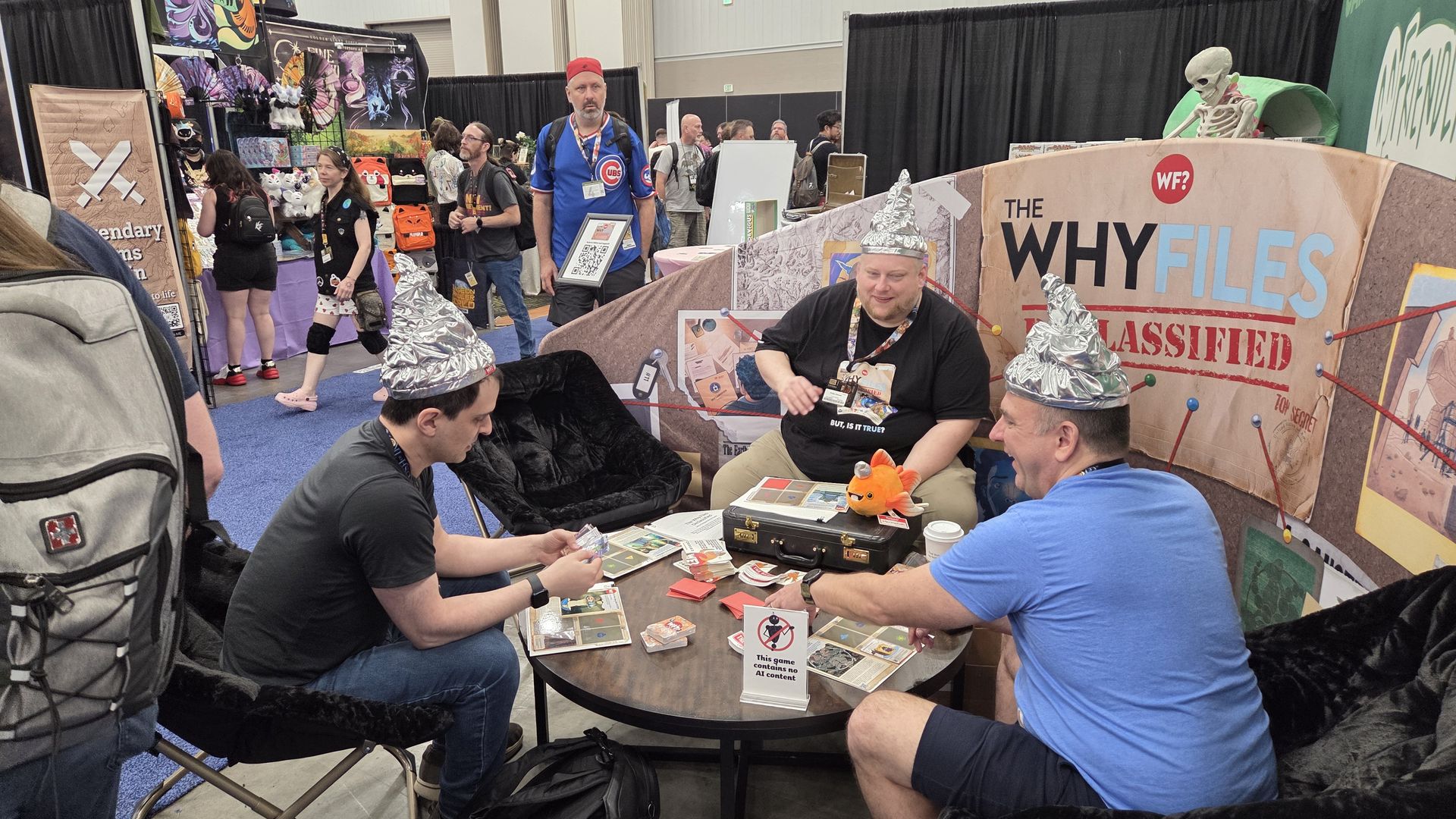 Convention scene: three men in aluminum foil hats play cards at a round table; behind them a booth reads "THE WHY FILES CLASSIFIED" with a skeleton prop, and other attendees wander the hall.