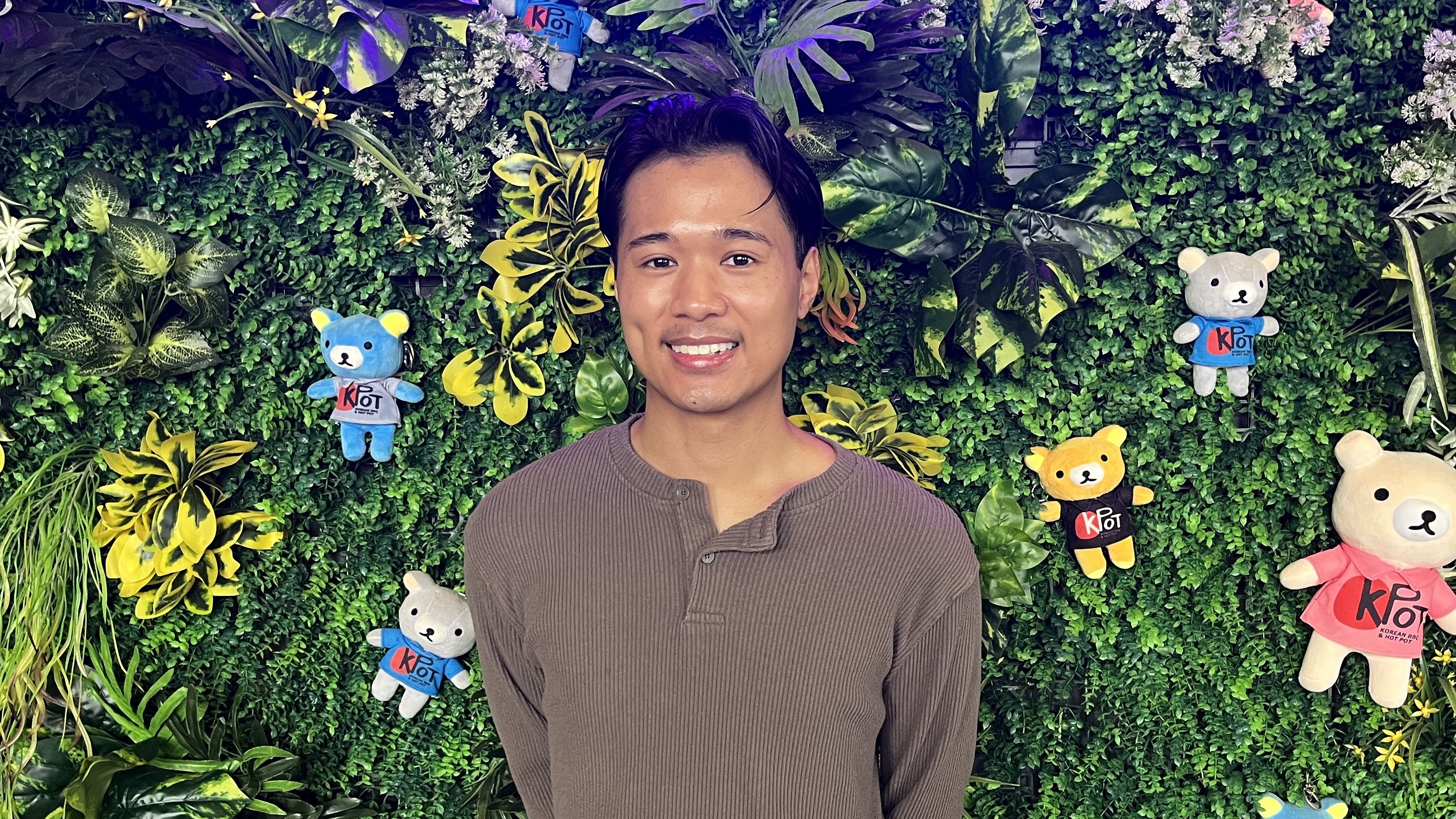 Jaye Xaymongkhonh, founder of Ethereal dance group, stands in front of a lush green wall decorated with various colorful plush bears wearing KPOT shirts.