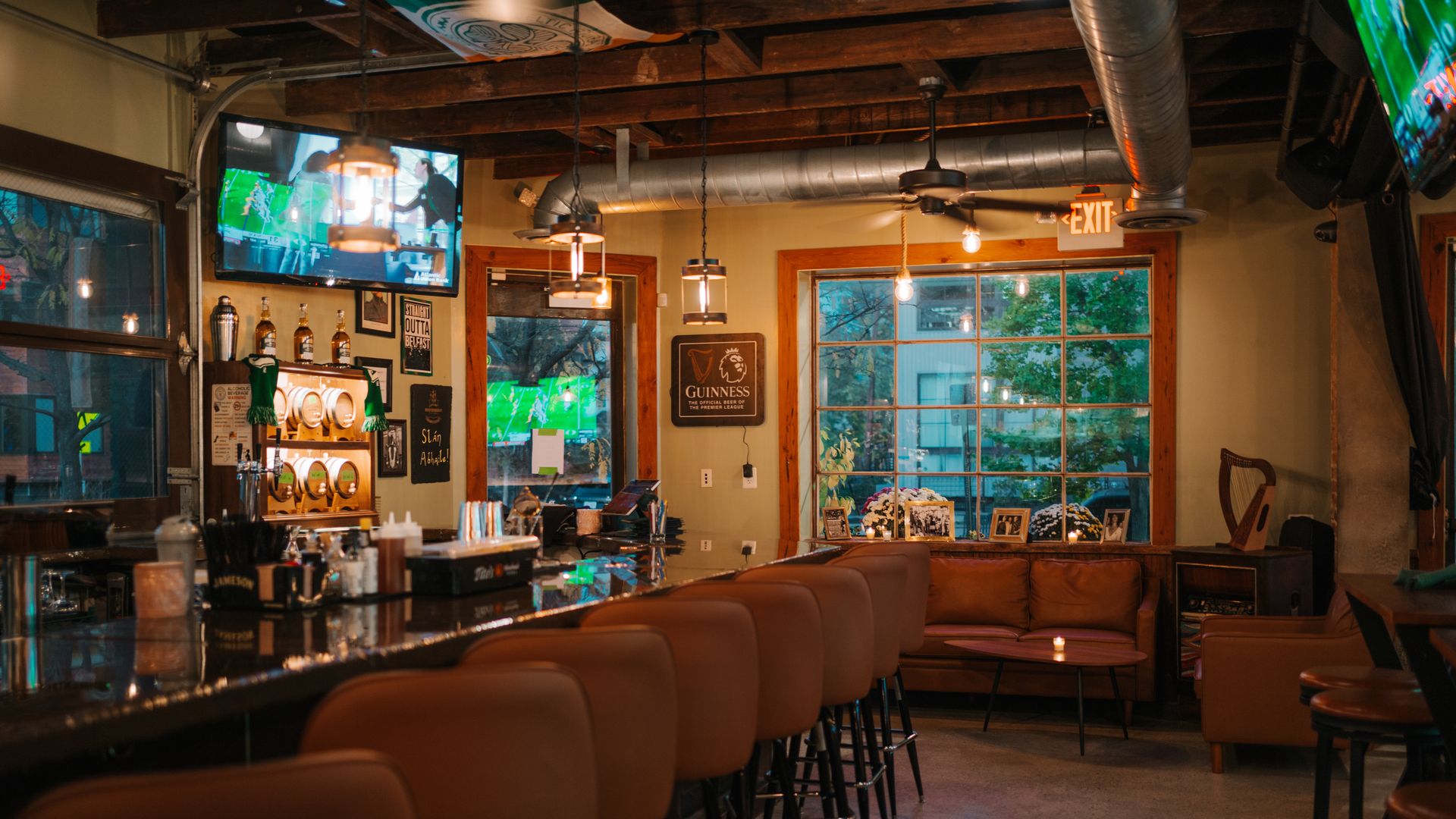 Cozy bar interior with brown leather stools lined along a dark counter, warm lighting, two TV screens showing sports, a large window, and a brown leather couch seating area.