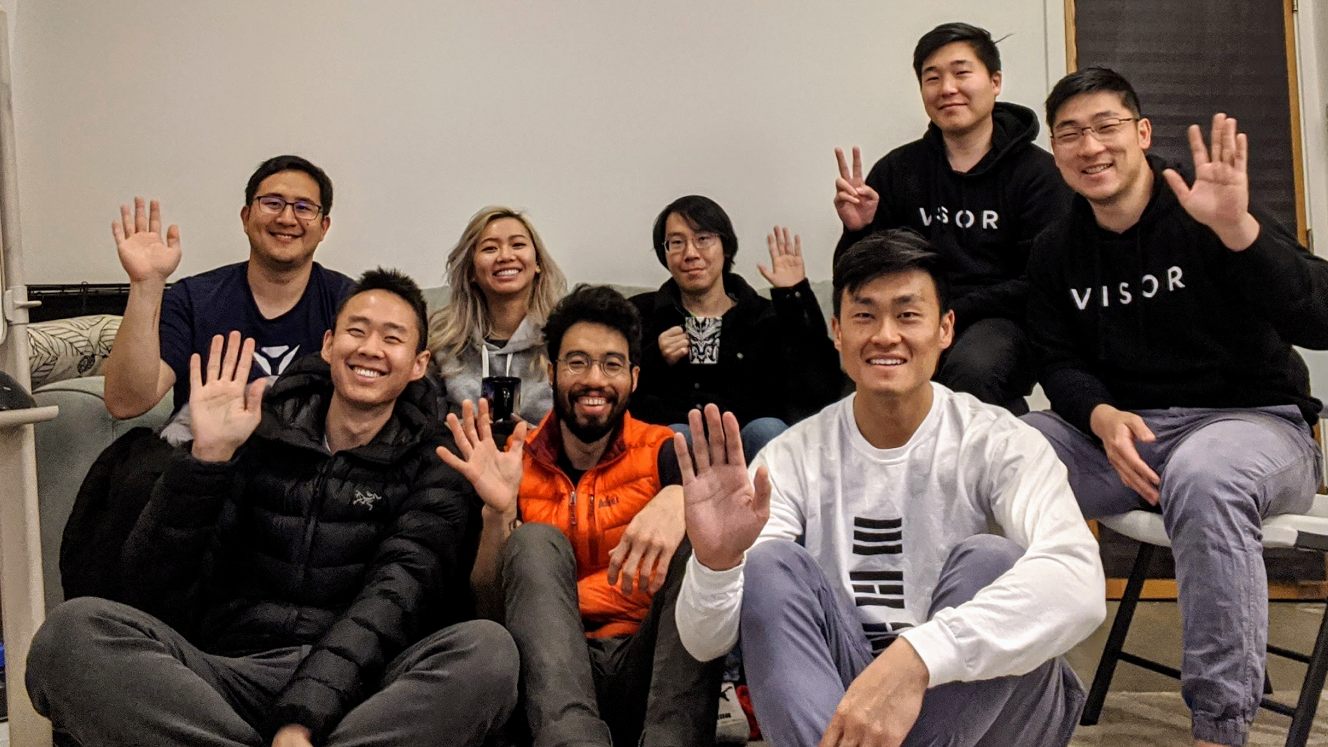 Eight people mostly wearing hoodies and jackets sit in a group without face masks and wave to the camera