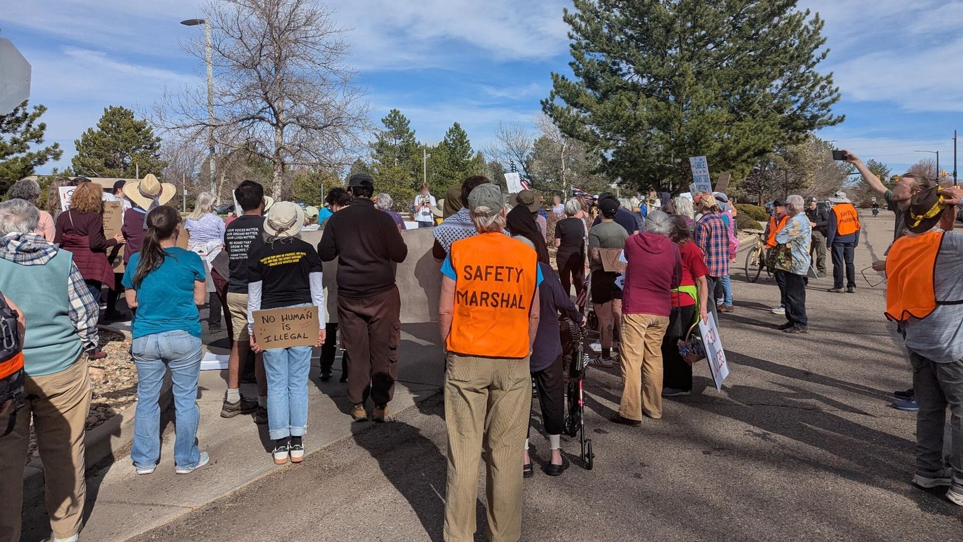 Group of people gathered outdoors for a protest, one holding a sign reading "NO HUMAN IS ILLEGAL," several wearing bright orange vests labeled "SAFETY MARSHAL," under a blue sky with trees.