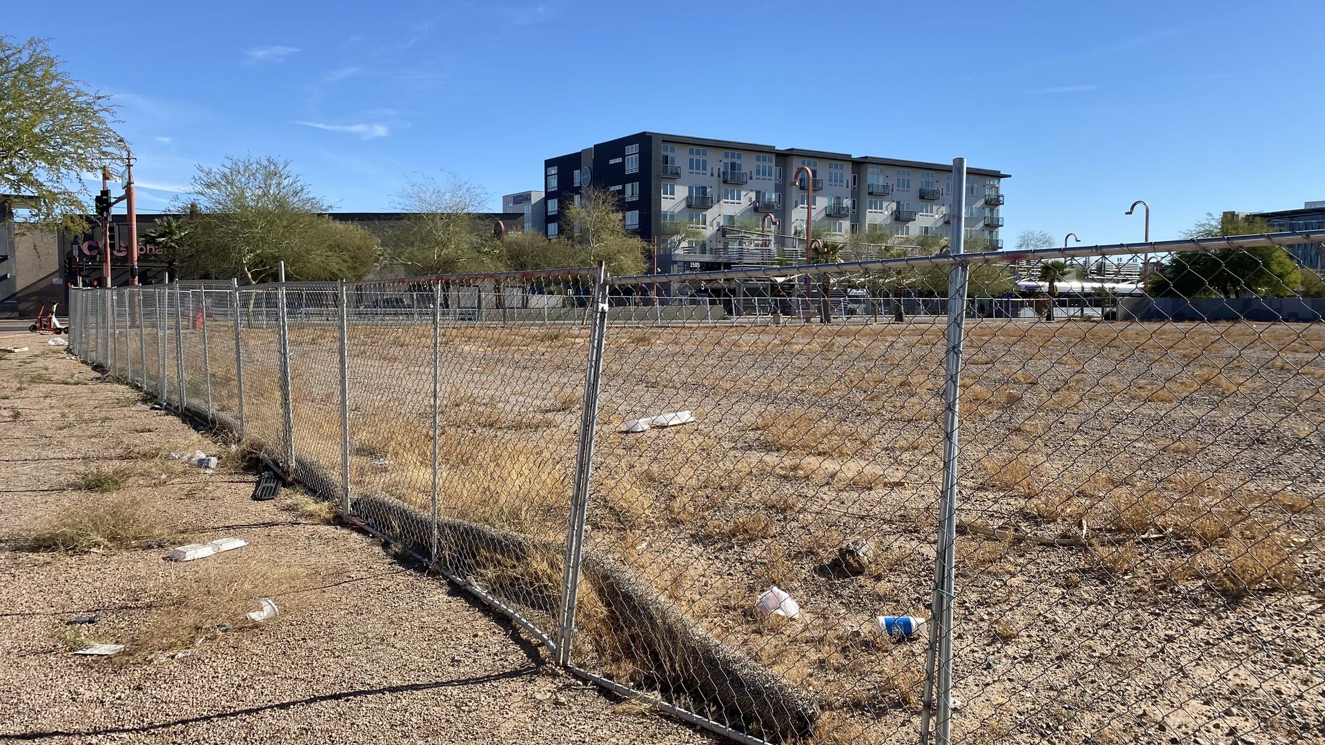 A  large dirt lot surrounded by a chain link fence with a luxury apartment building in the background.