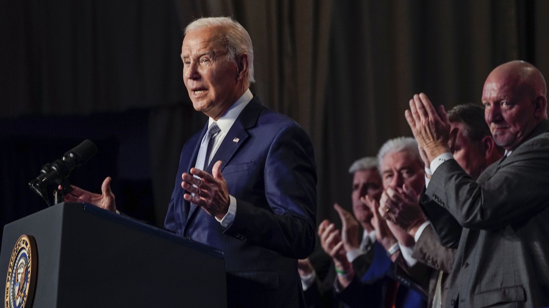 Joe Biden stands at a podium, speaking to a crowd, while a line of people clap behind him.