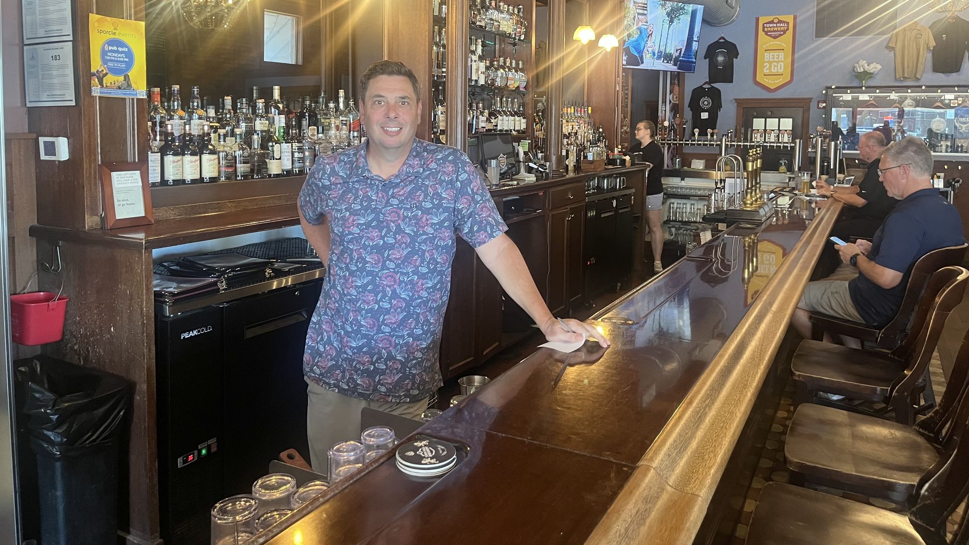 Pete Rifakes in a blue floral shirt smiling behind a wooden bar counter with bottles and glasses. Two men sitting at the bar and a woman working in the background under warm hanging lights.