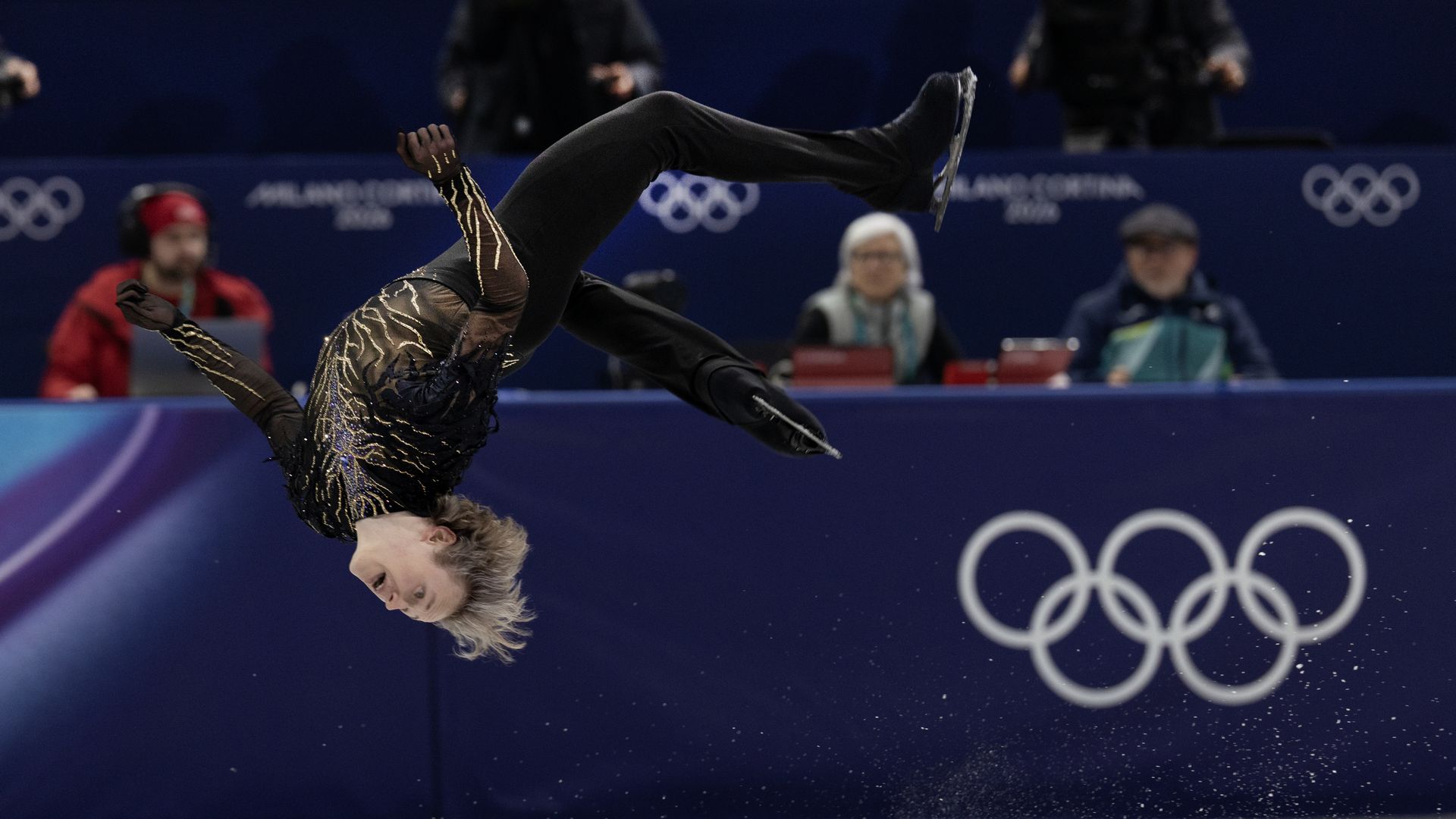 A male figure skater in mid-air about to do a backflip on the ice. There's the Olympics logo to his right.