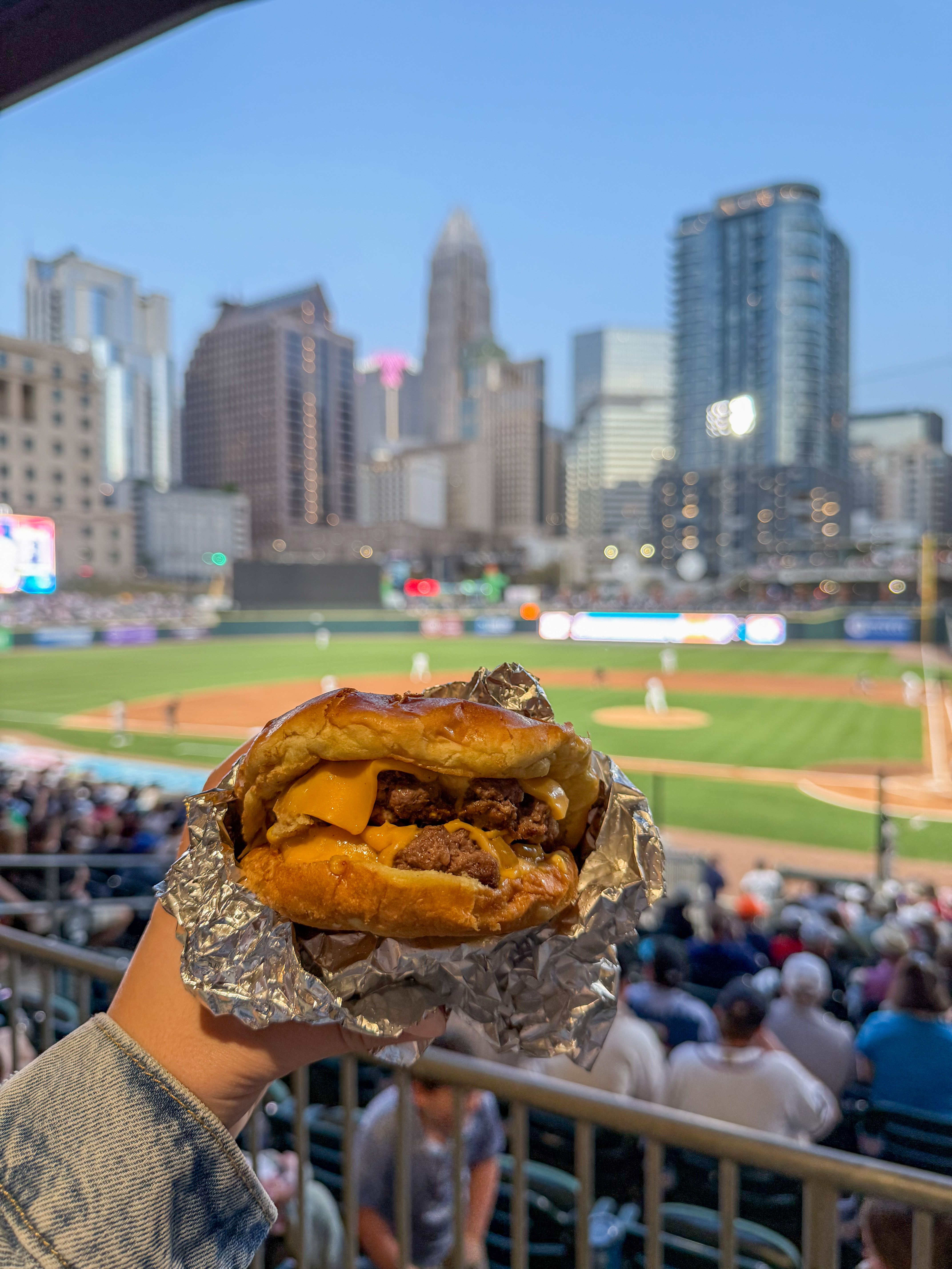 A hand holds a juicy cheeseburger wrapped in foil with the baseball field and city skyline in the background.