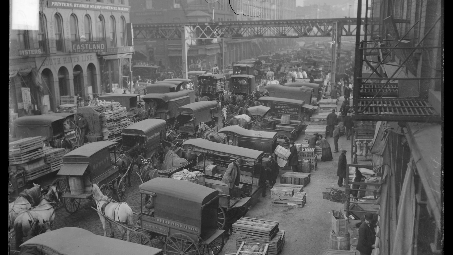 Black-and-white street scene from the early 20th century: crowded cobbled road with horse-drawn wagons, crates, and a few trucks, storefronts with signs and an elevated iron bridge overhead.