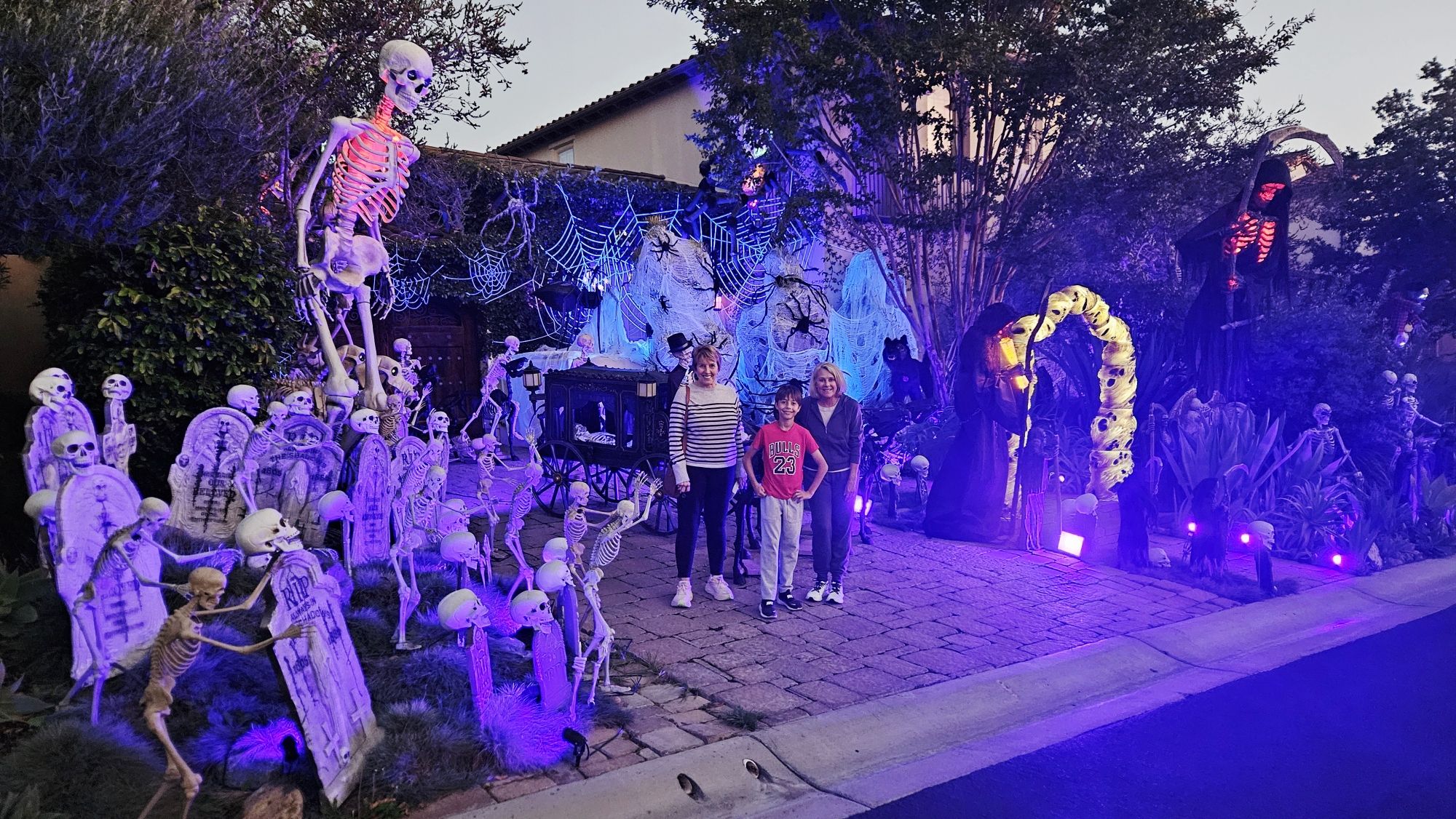 Three people stand smiling in a Halloween-decorated yard with skeletons, tombstones, giant spider webs, and grim reaper figures lit by purple and orange lights at dusk.