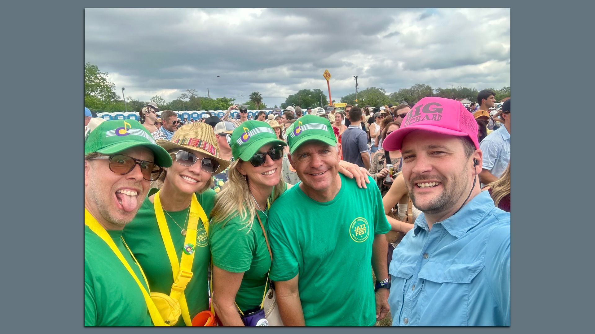 Selfie at a music festival. Foreground group in green shirts and hats with a music-note logo; a woman in sunglasses and straw hat; a man in a bright pink cap. Crowd, porta-potties, and cloudy sky in back.