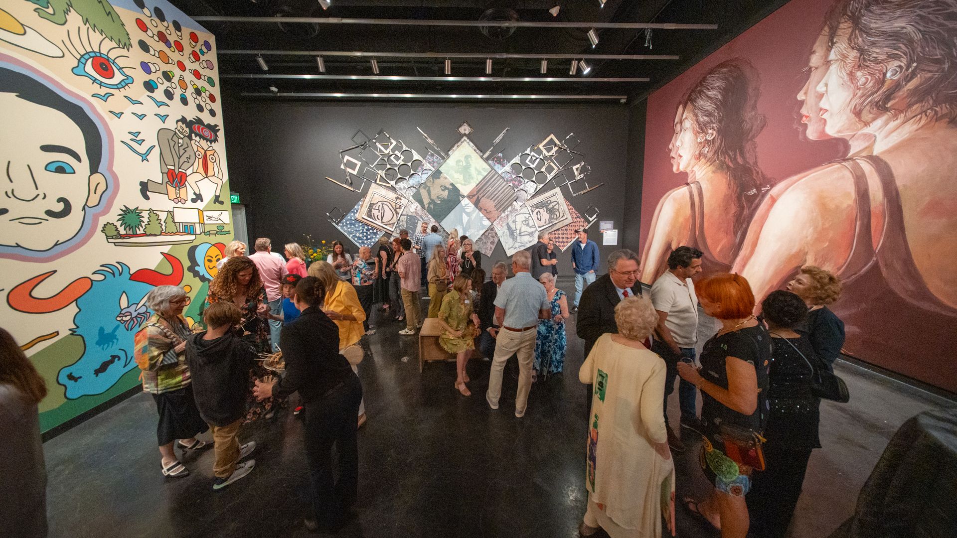 People gathered in an art gallery with colorful murals on the walls, including a large face with whimsical elements on the left and two women in profile on the right.