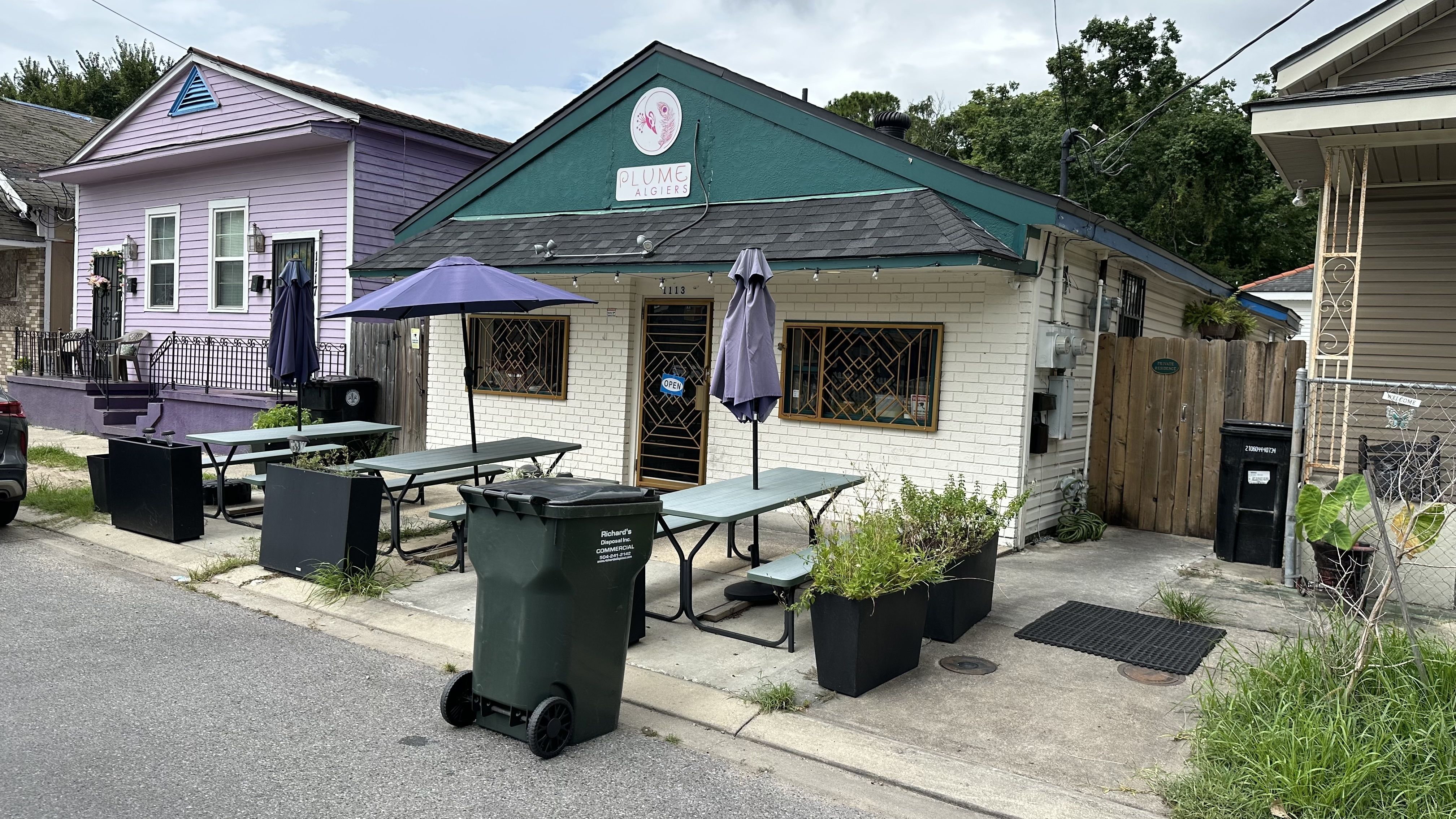 Street view of a small restaurant with green roof and white brick walls, outdoor green tables with purple umbrellas, large green trash bin, and a purple house beside it.