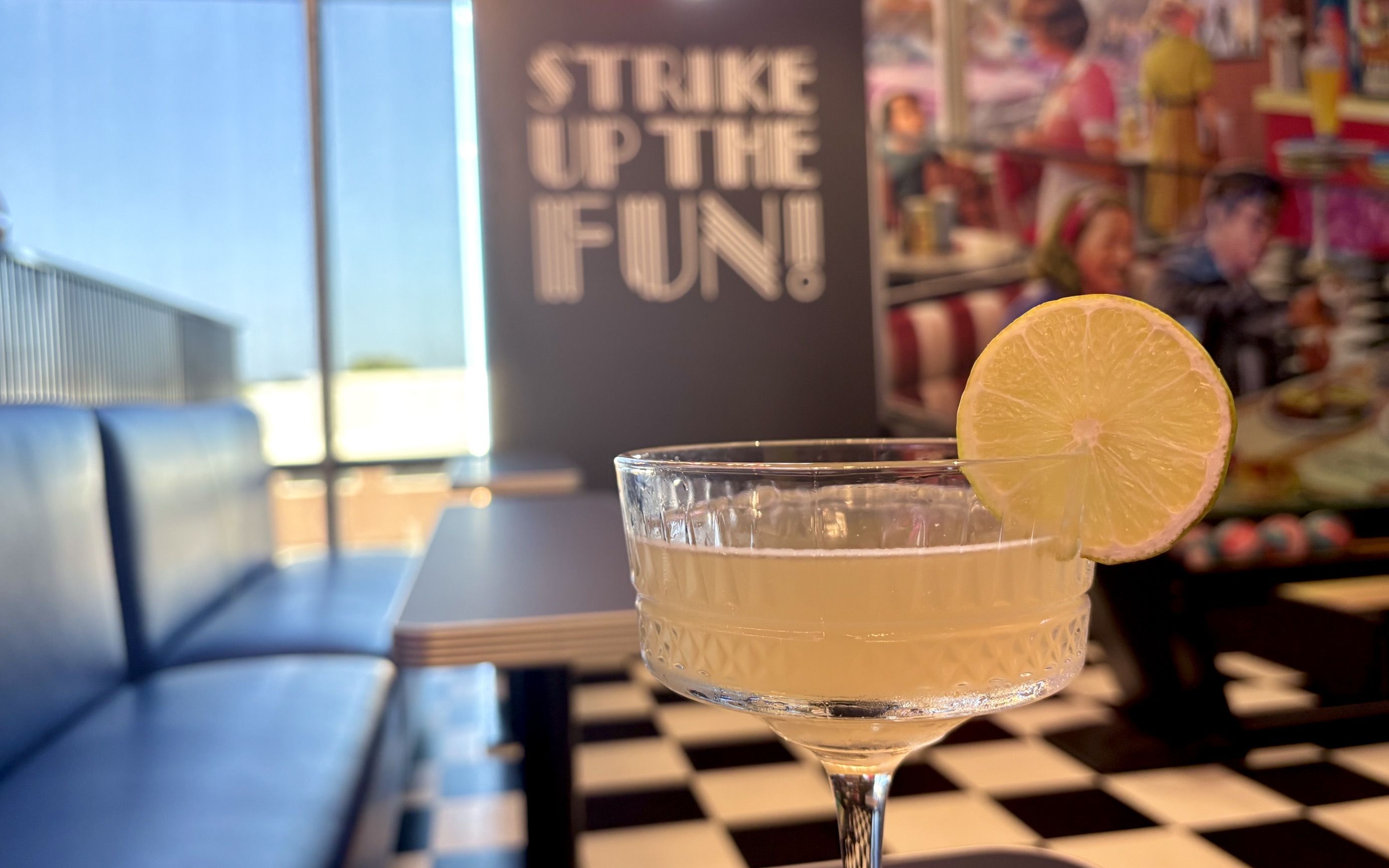 Close-up of a lemon-garnished cocktail in a crystal stem glass on a bar, with a blurred retro diner backdrop and a sign reading "STRIKE UP THE FUN!" amid blue seating.