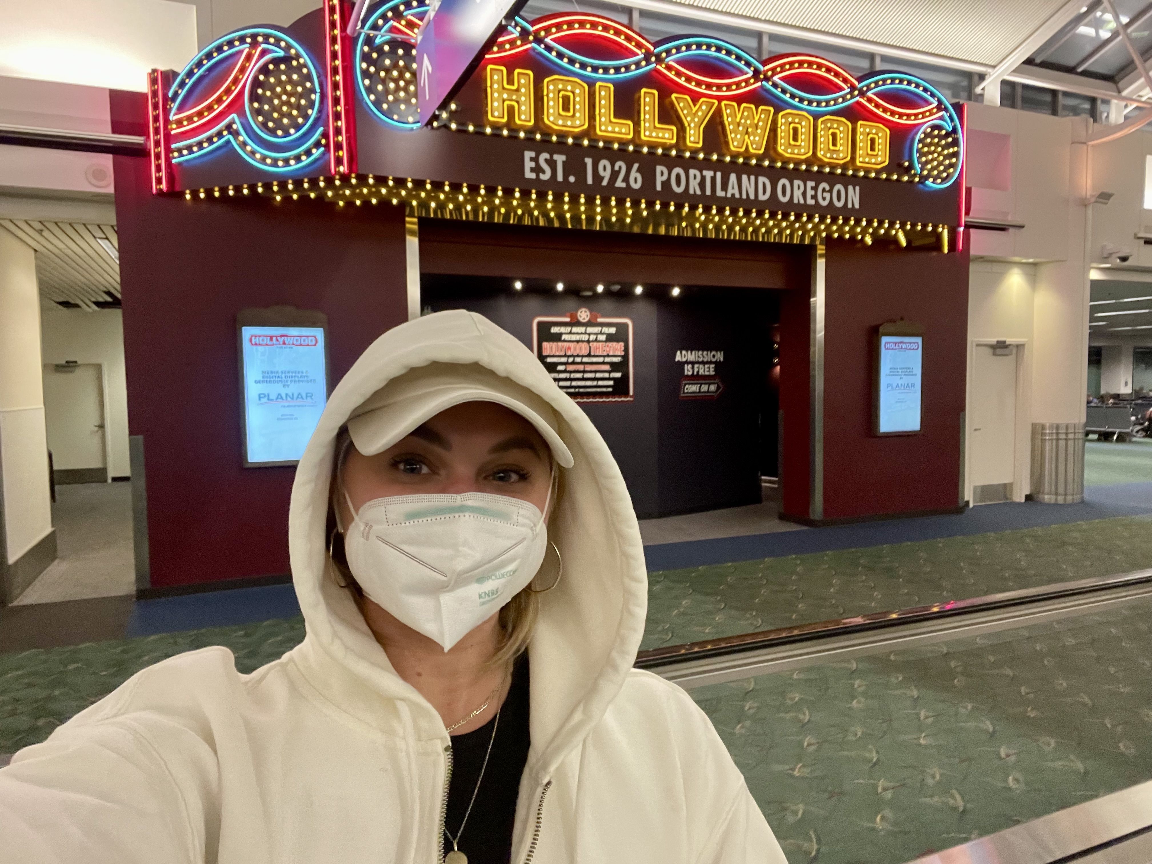 Person wearing a white hooded jacket and face mask takes selfie in front of colorful illuminated HOLLYWOOD sign inside a building, with green carpeted floor and signs indicating free admission.